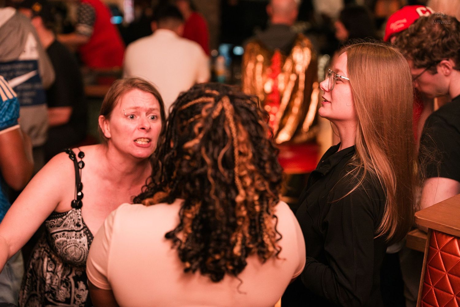 Three women engaged in conversation at a dimly lit bar or nightclub gathering.