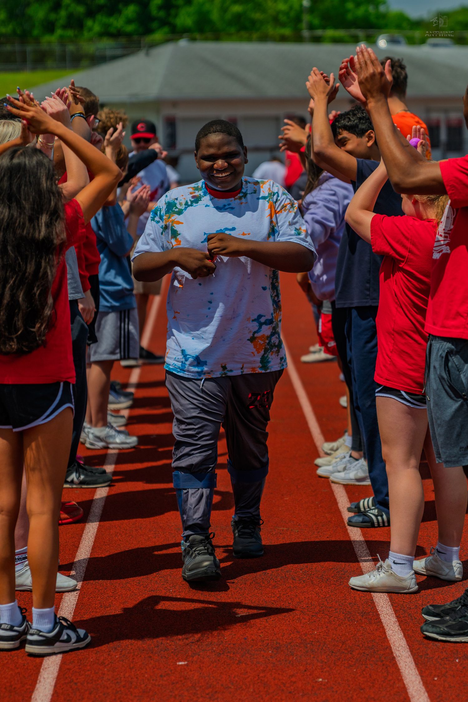 Student walks through a celebratory high-five tunnel on a red track during an outdoor school sports event on a sunny day.