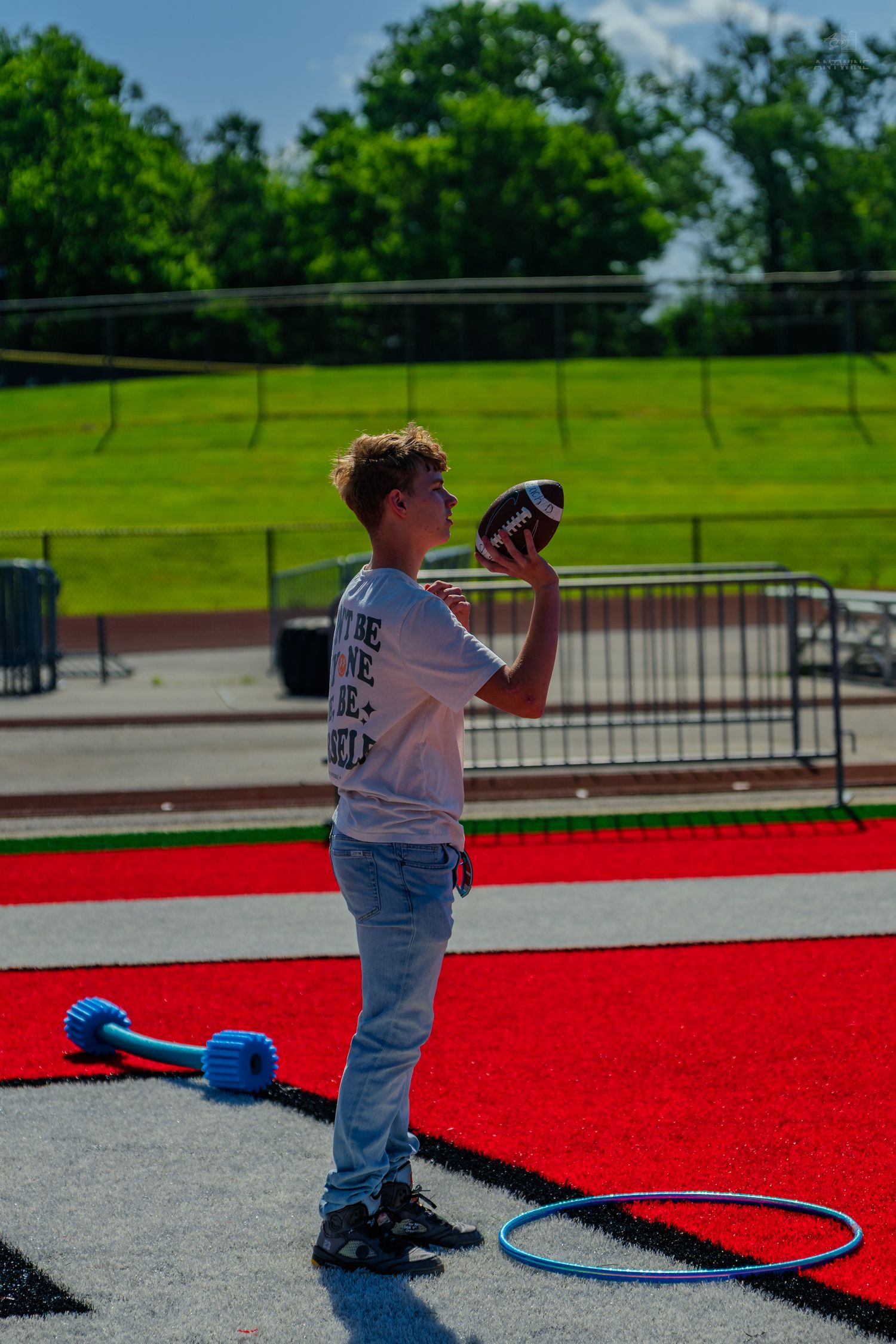 A teenage boy holds a football on a red turf field with bleachers and green trees in the background.