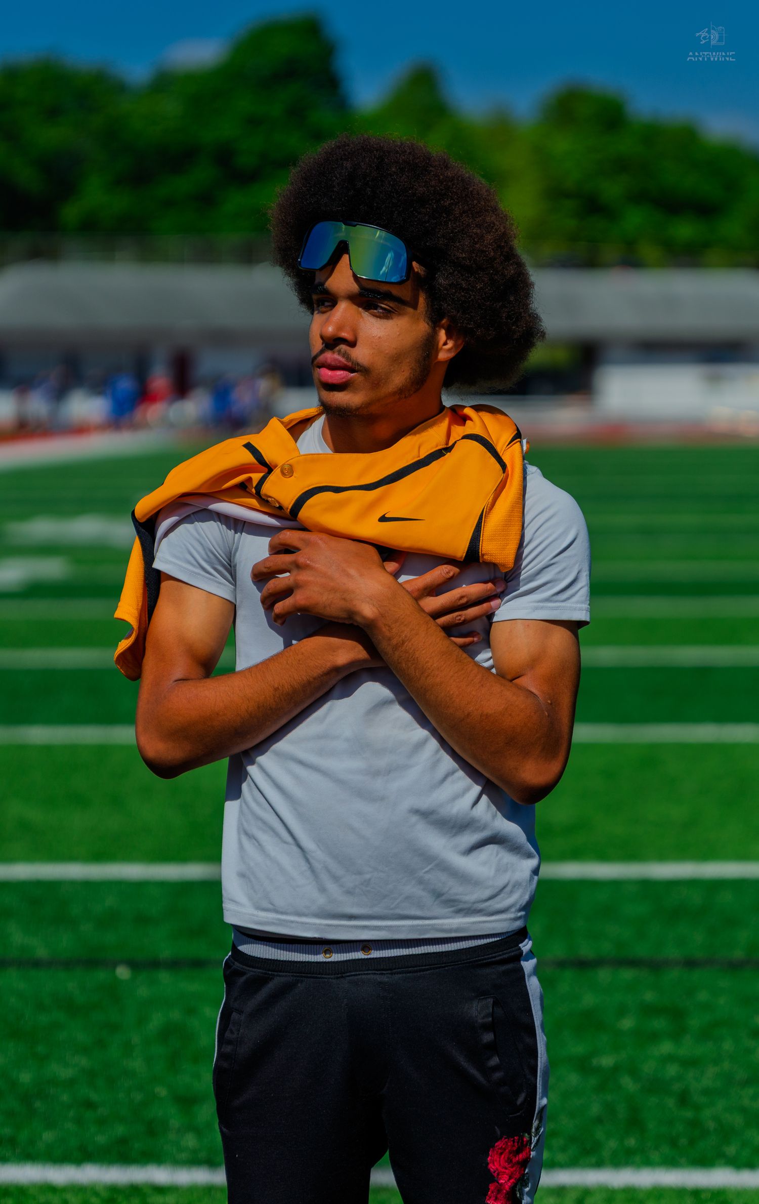 Young athlete in gray Nike shirt and sunglasses holds orange jersey on a green football field.