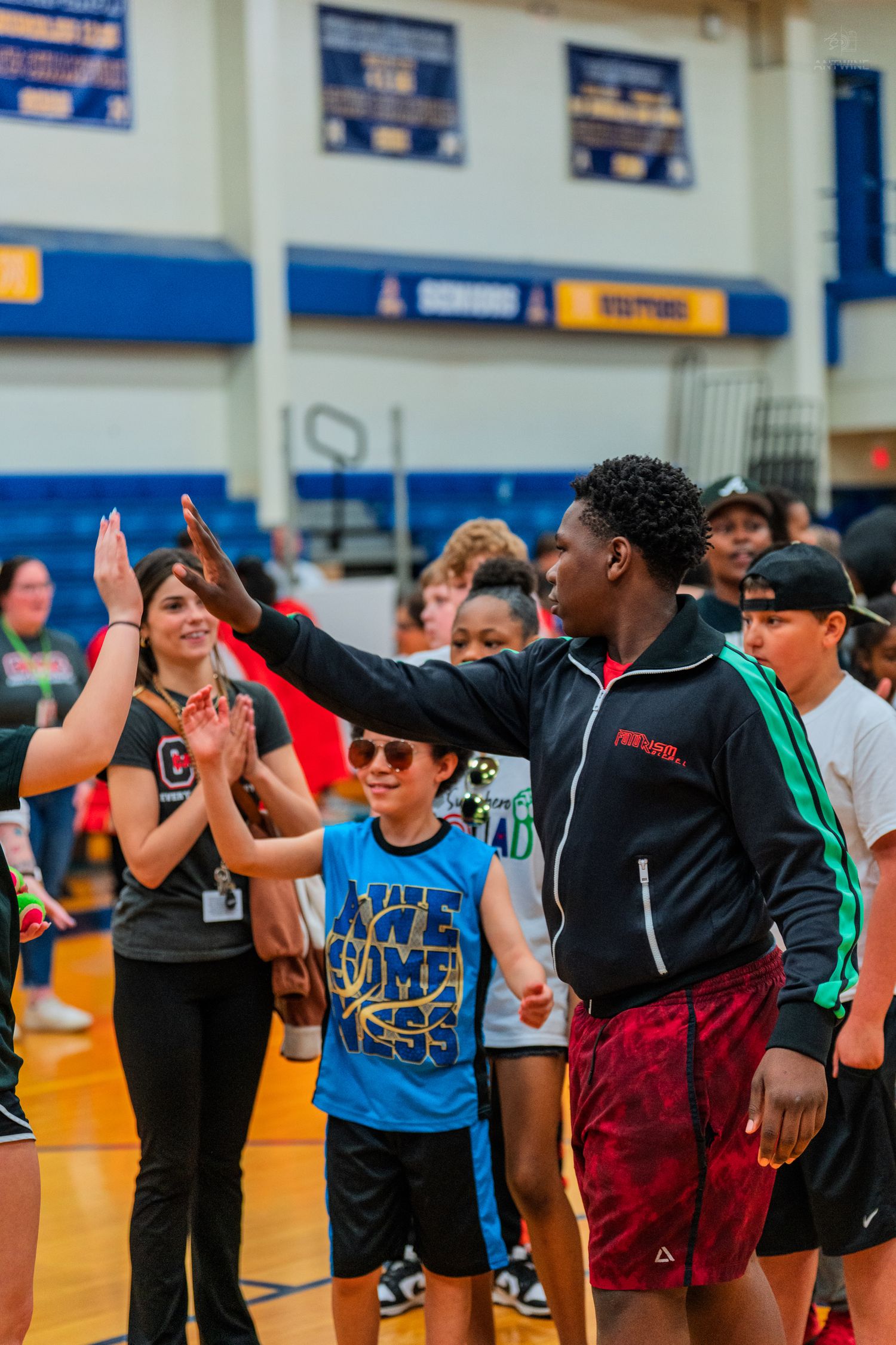 Tall teen gives high-five to younger student in blue shirt at school gymnasium event with crowd in background.