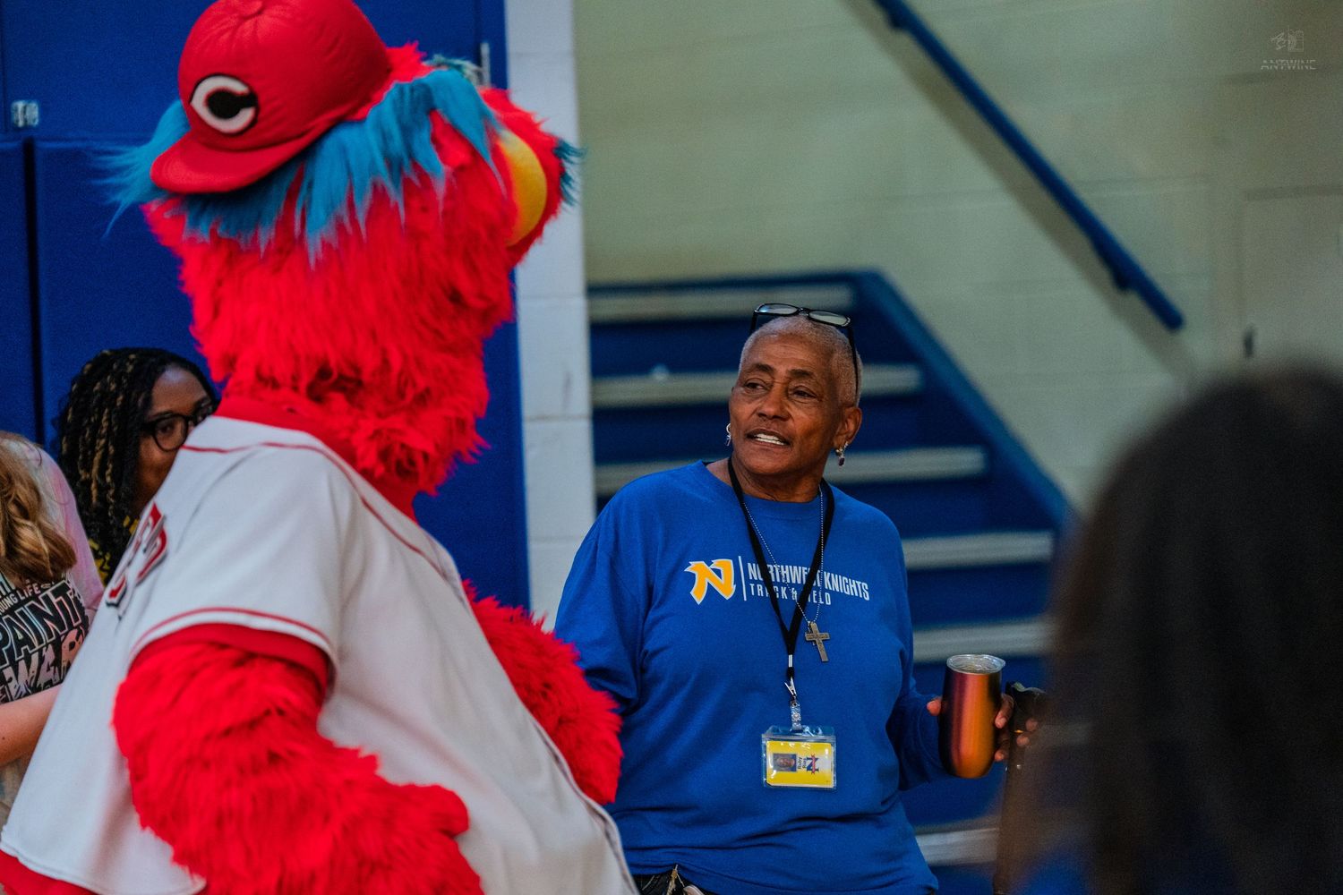 A smiling woman in a blue shirt laughs with a colorful red bird mascot at an indoor school event.