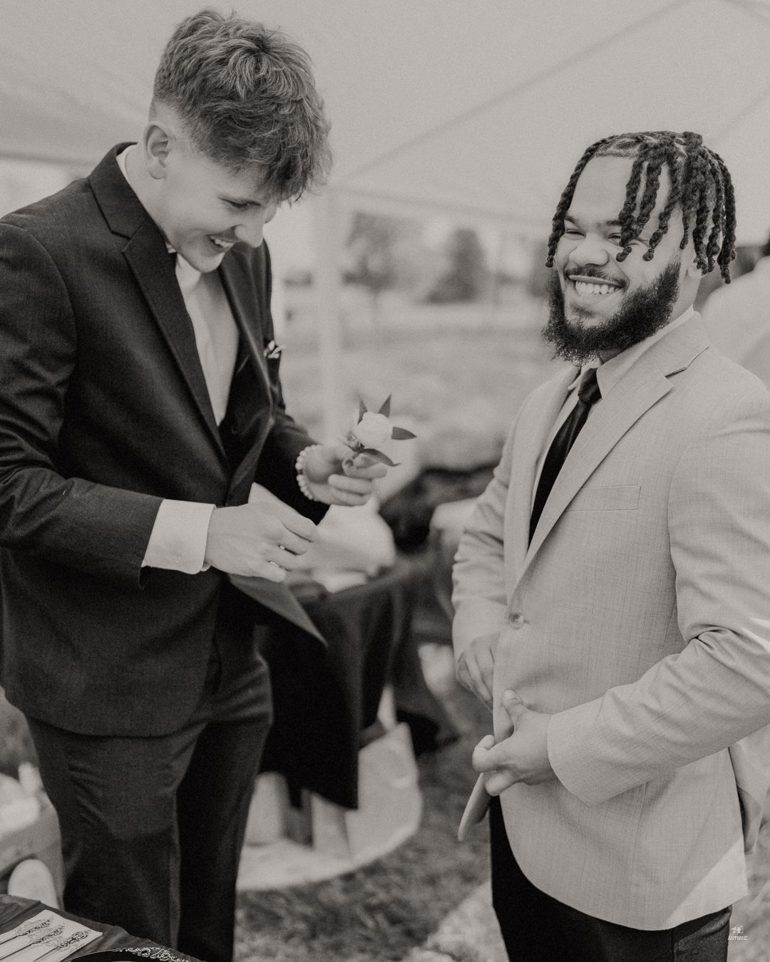 Black and white photo of two wedding guests in suits laughing and talking under a tent.