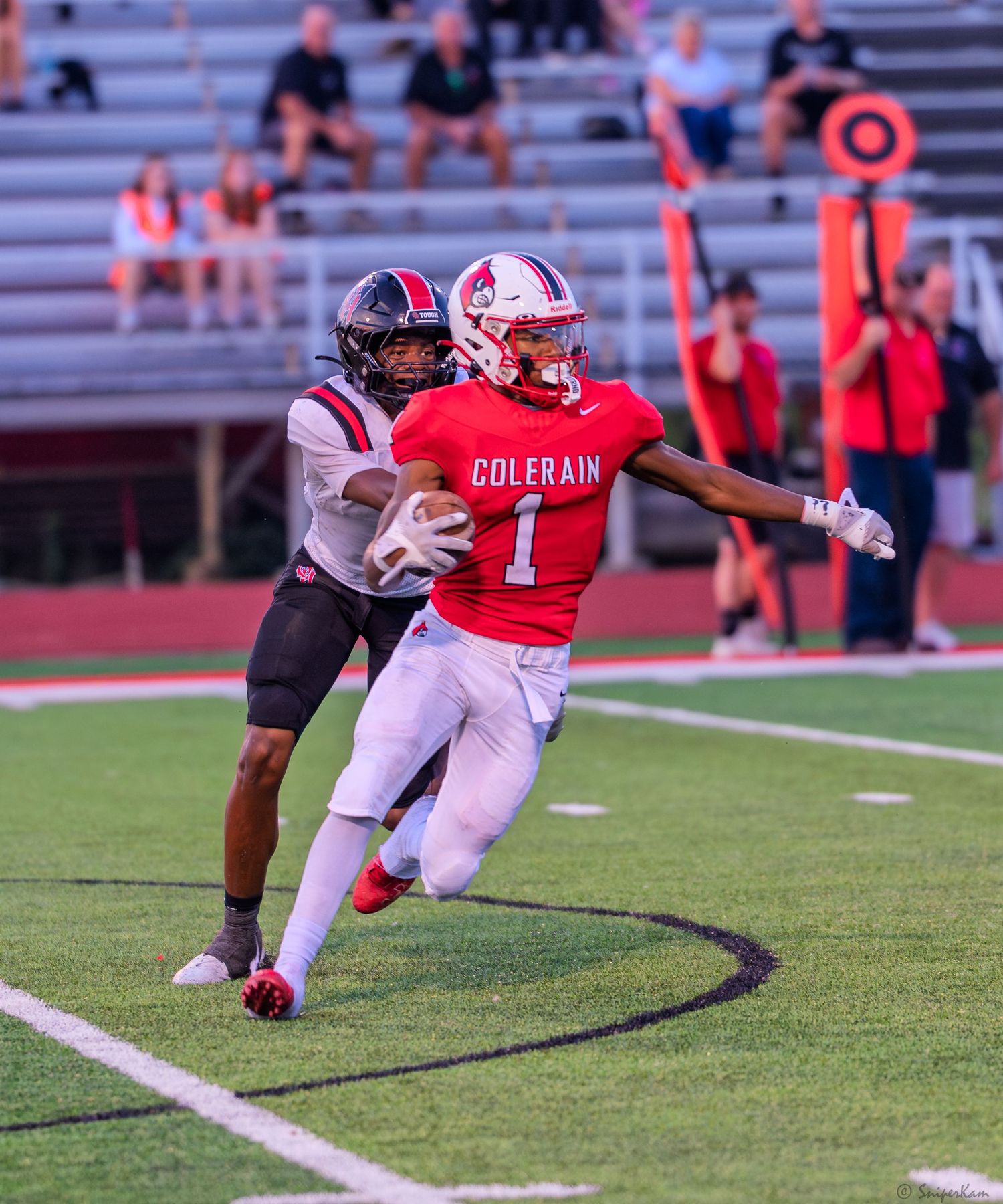 Colerain #1 receiver in red uniform runs along the sideline after a catch with a defender in pursuit during a football game.