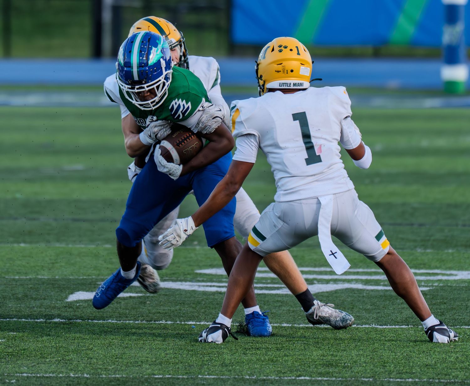 A ball carrier in blue and green uniform pushes through defenders in white and gold during a high school game.
