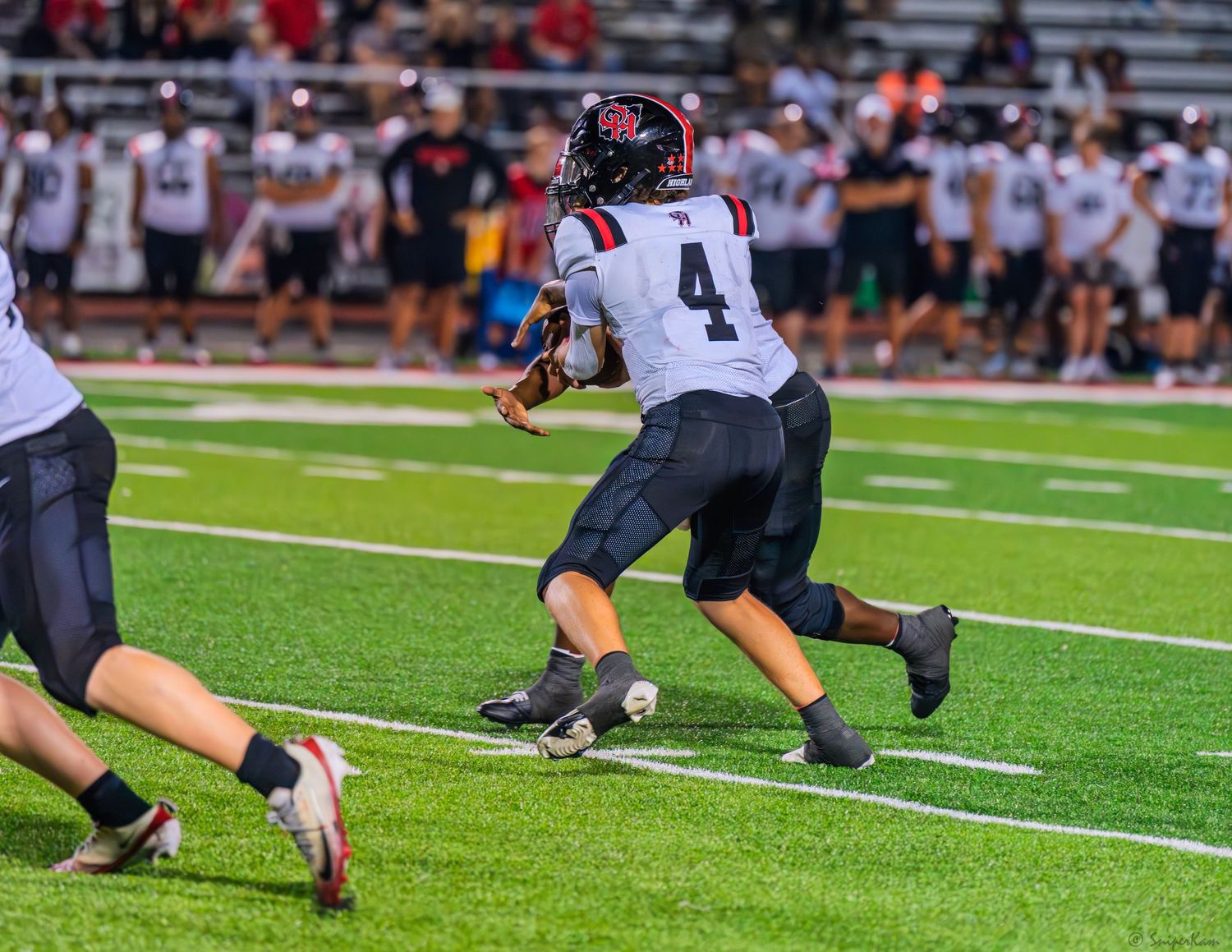 High school football player number 4 in dark uniform carries the ball during a night game.