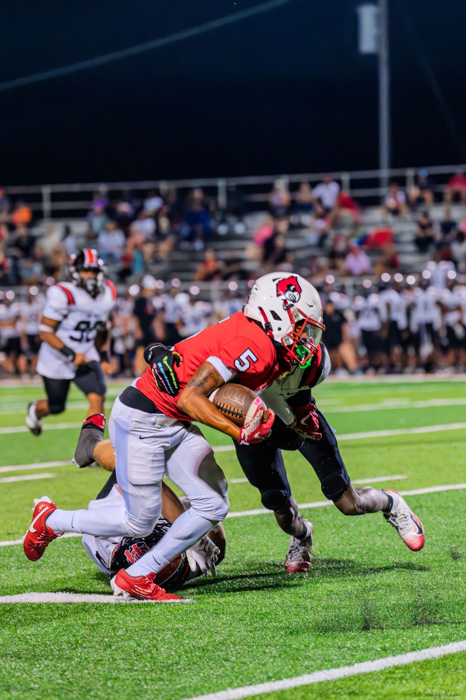 A red-uniformed ball carrier #5 fights through a tackle attempt while driving forward during a high school football game.
