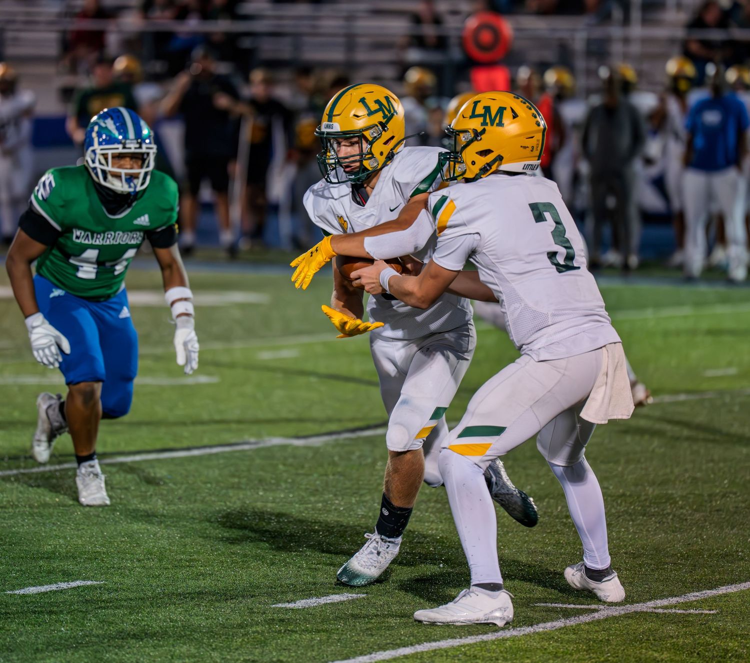 High school football quarterback hands off to number 3 receiver while blue-uniformed defender pursues the play.