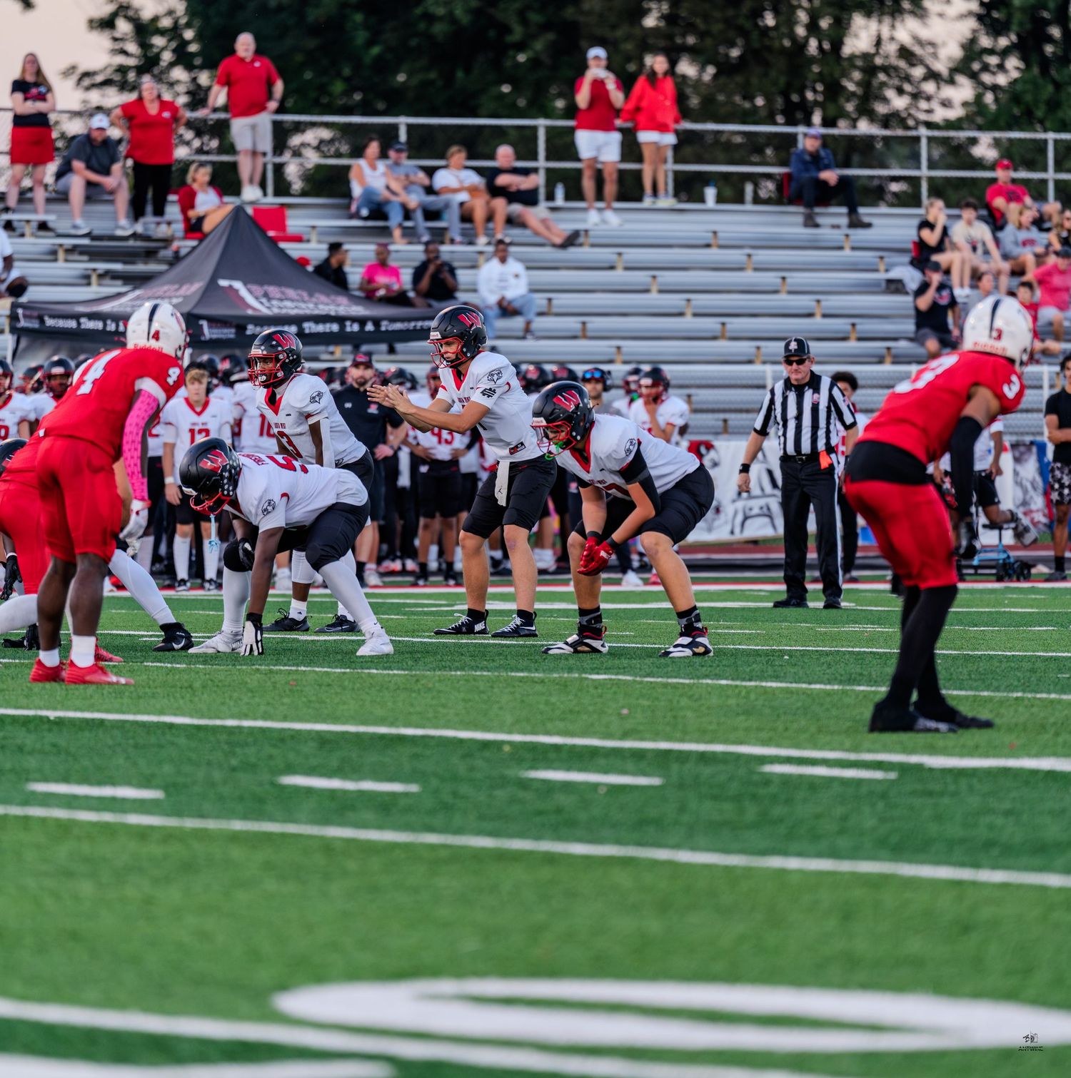 White-uniformed football team lines up against red-uniformed opponents on a green field during a high school game.