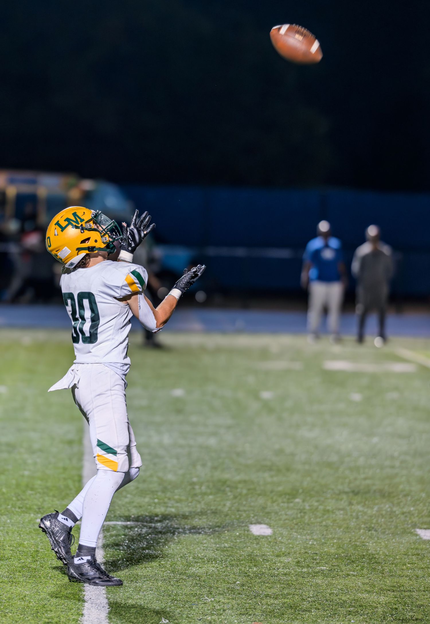 Youth football player #30 in green and gold uniform reaches up to catch a football during a night game.