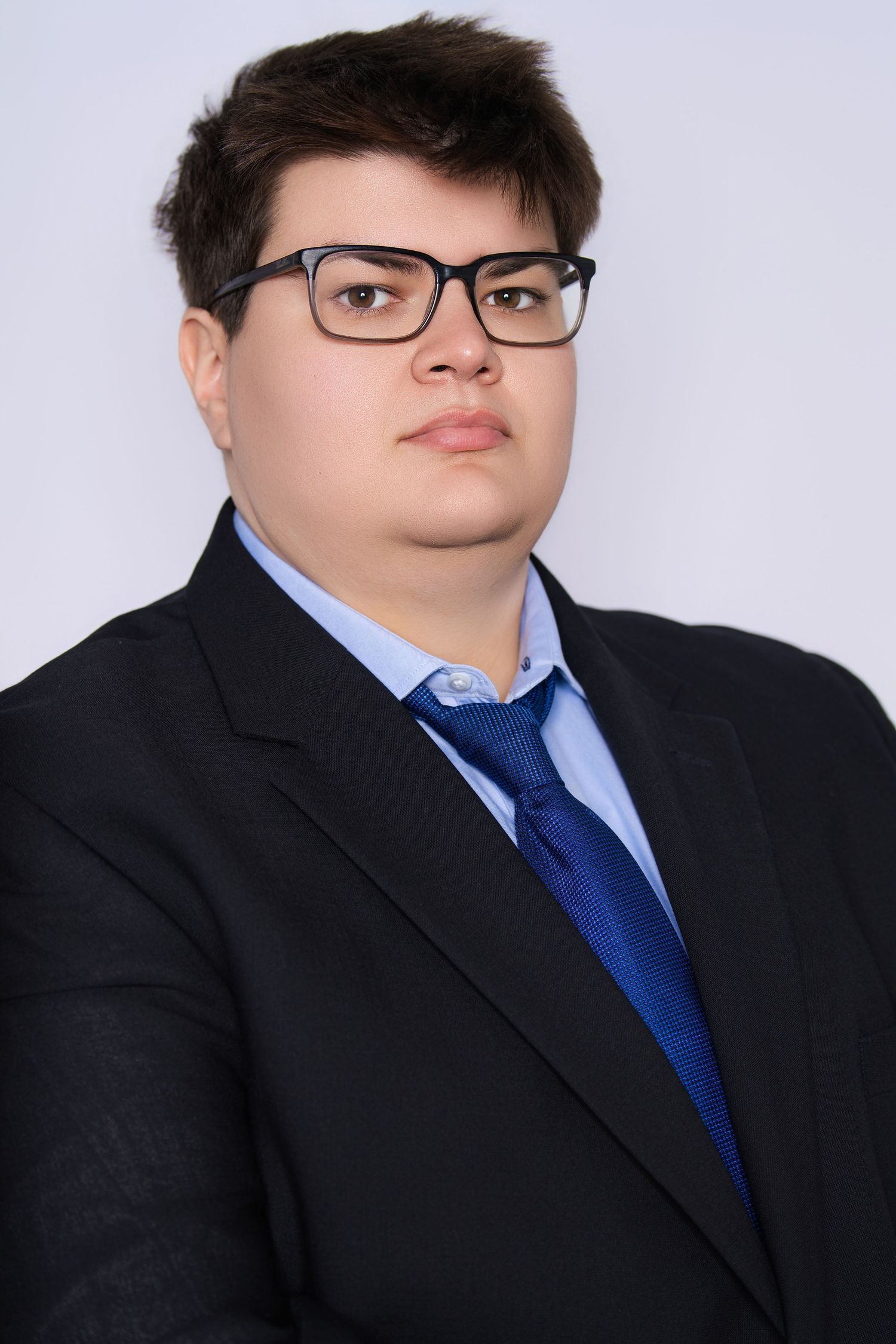 Young man in black suit, blue tie, and glasses poses for a professional portrait photo.