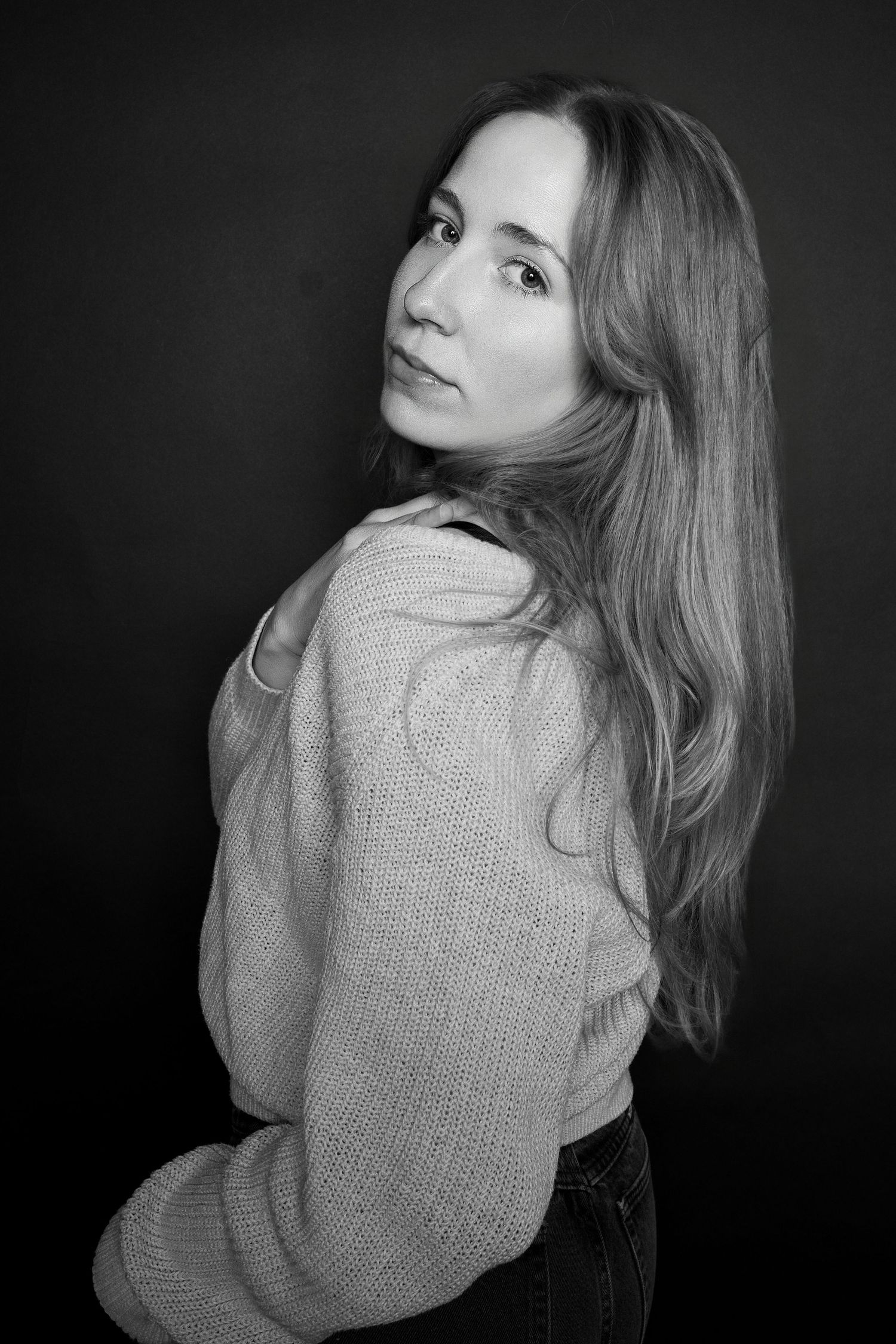 Black and white portrait of a woman with long hair in a knit sweater, looking over her shoulder.