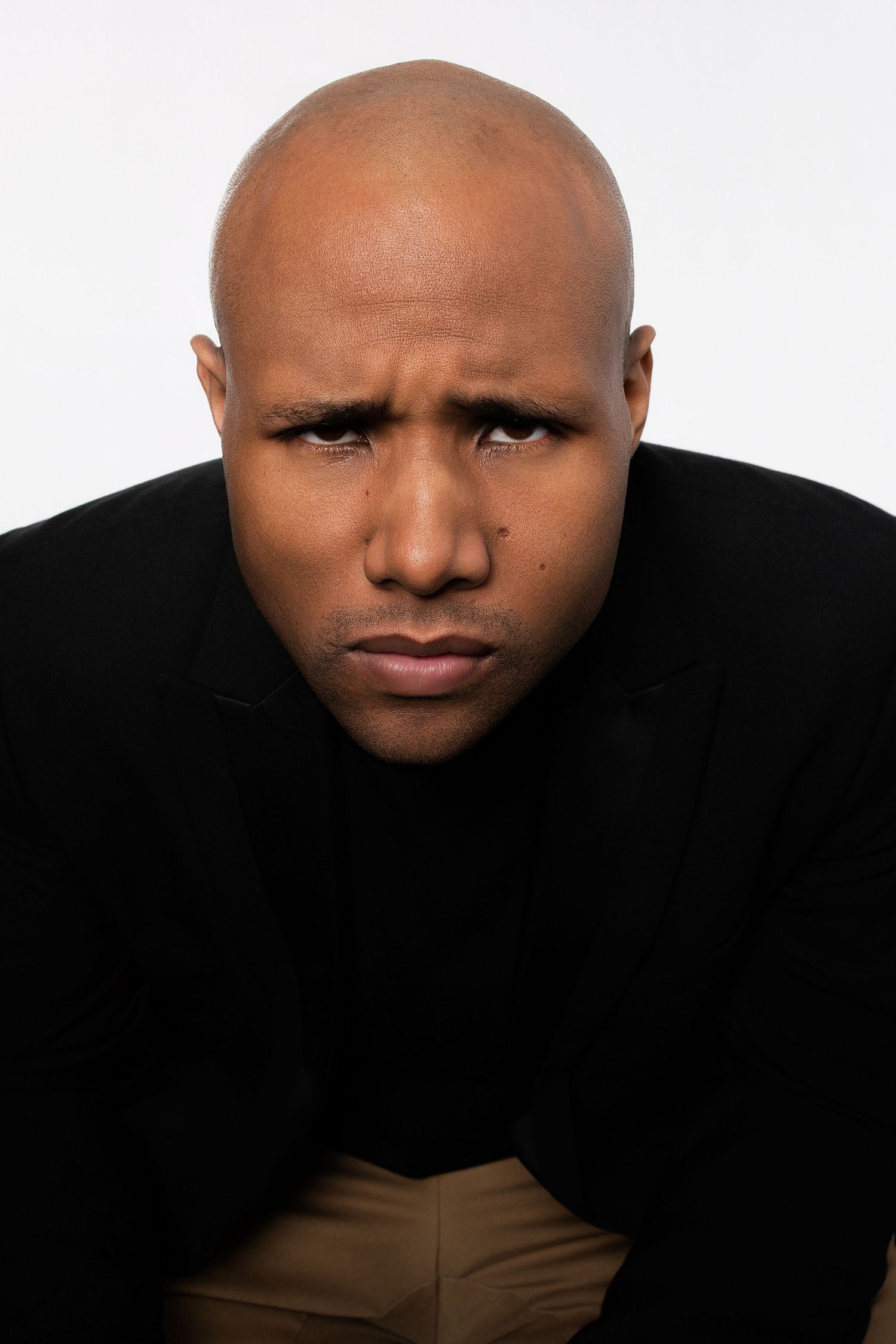 Bald Black man in black blazer leaning forward with stern frowning expression in studio photo.