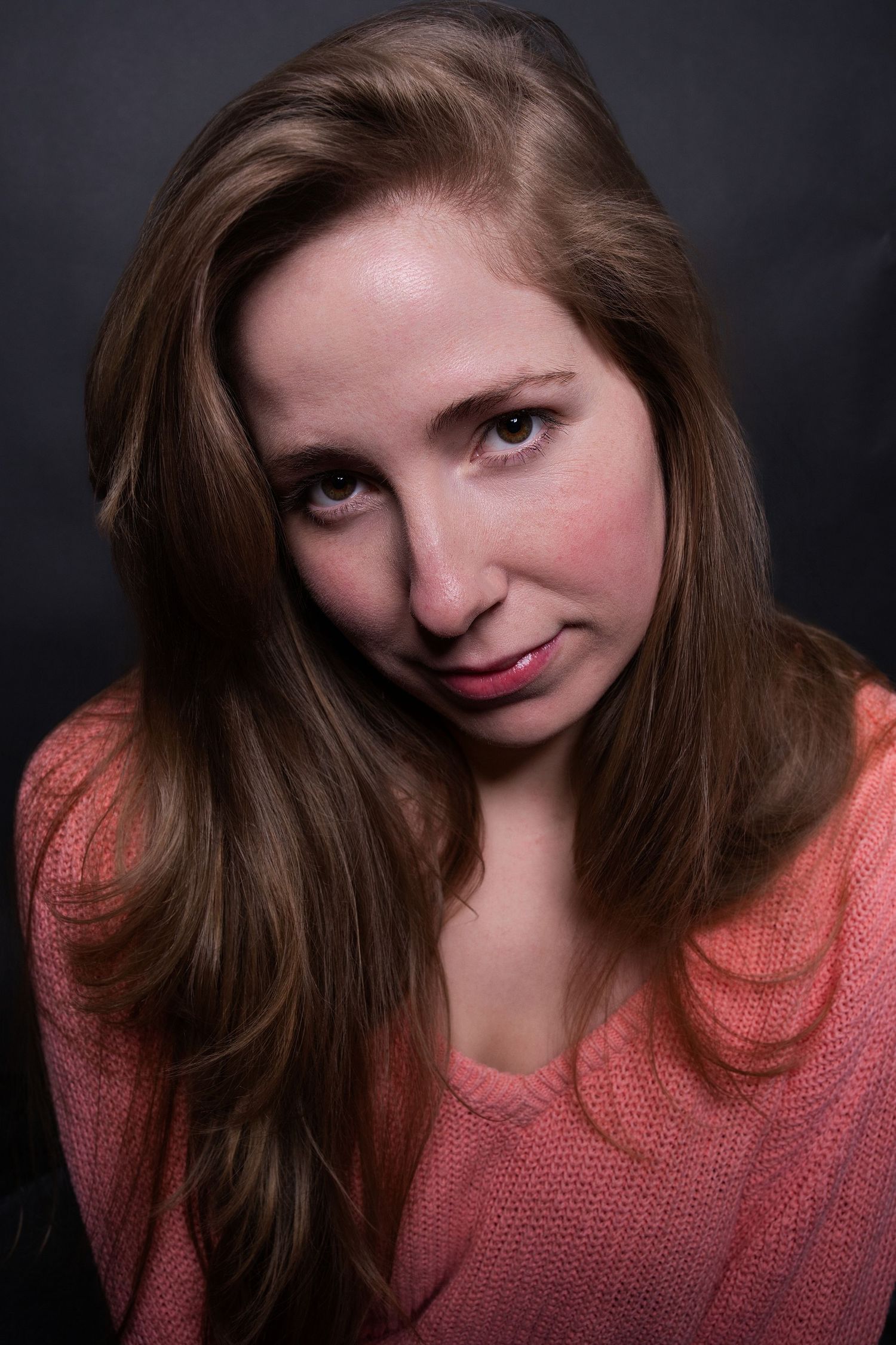 Young woman with long brown hair wearing a pink knit sweater poses for a studio portrait against dark background.