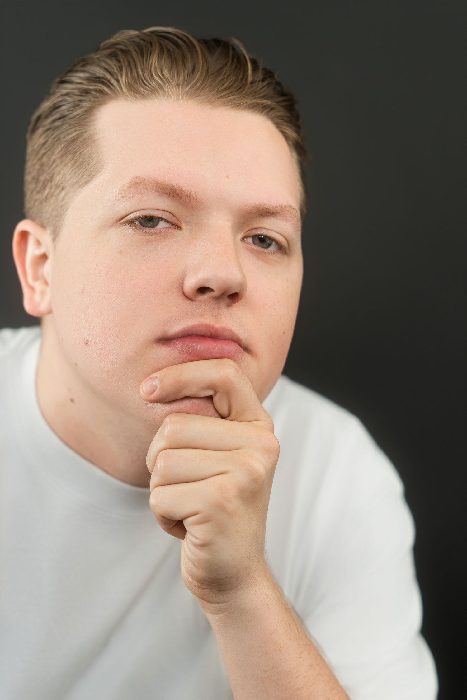 Young man in white t-shirt resting chin on fist, thoughtful expression against dark background.