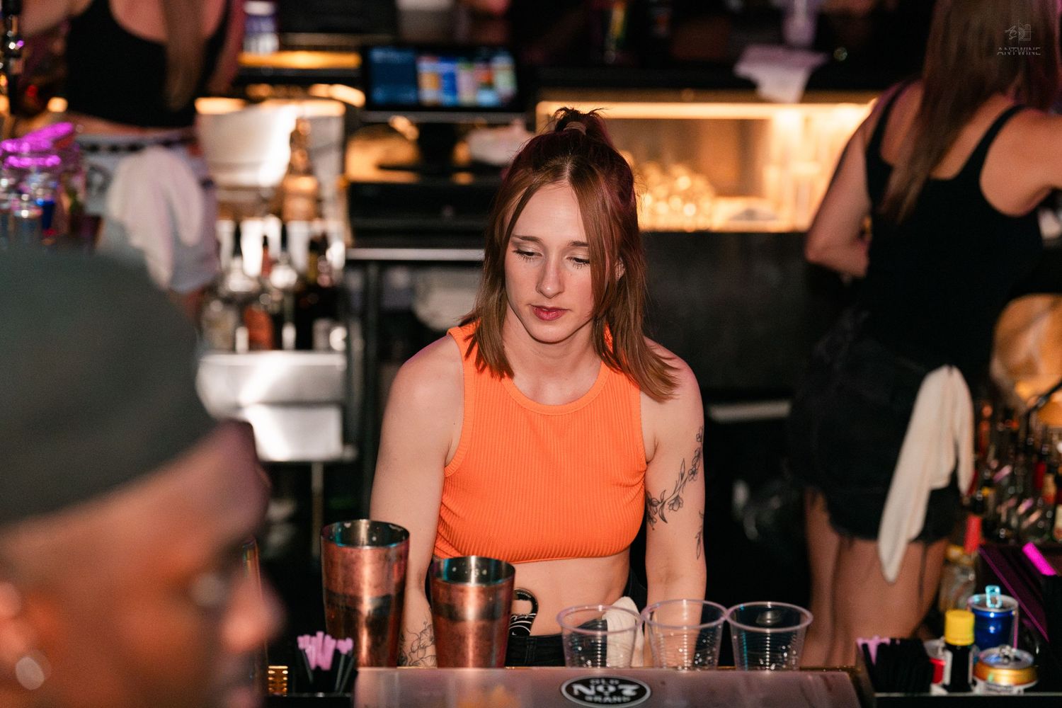 Female bartender in orange crop top prepares cocktails at a busy bar.