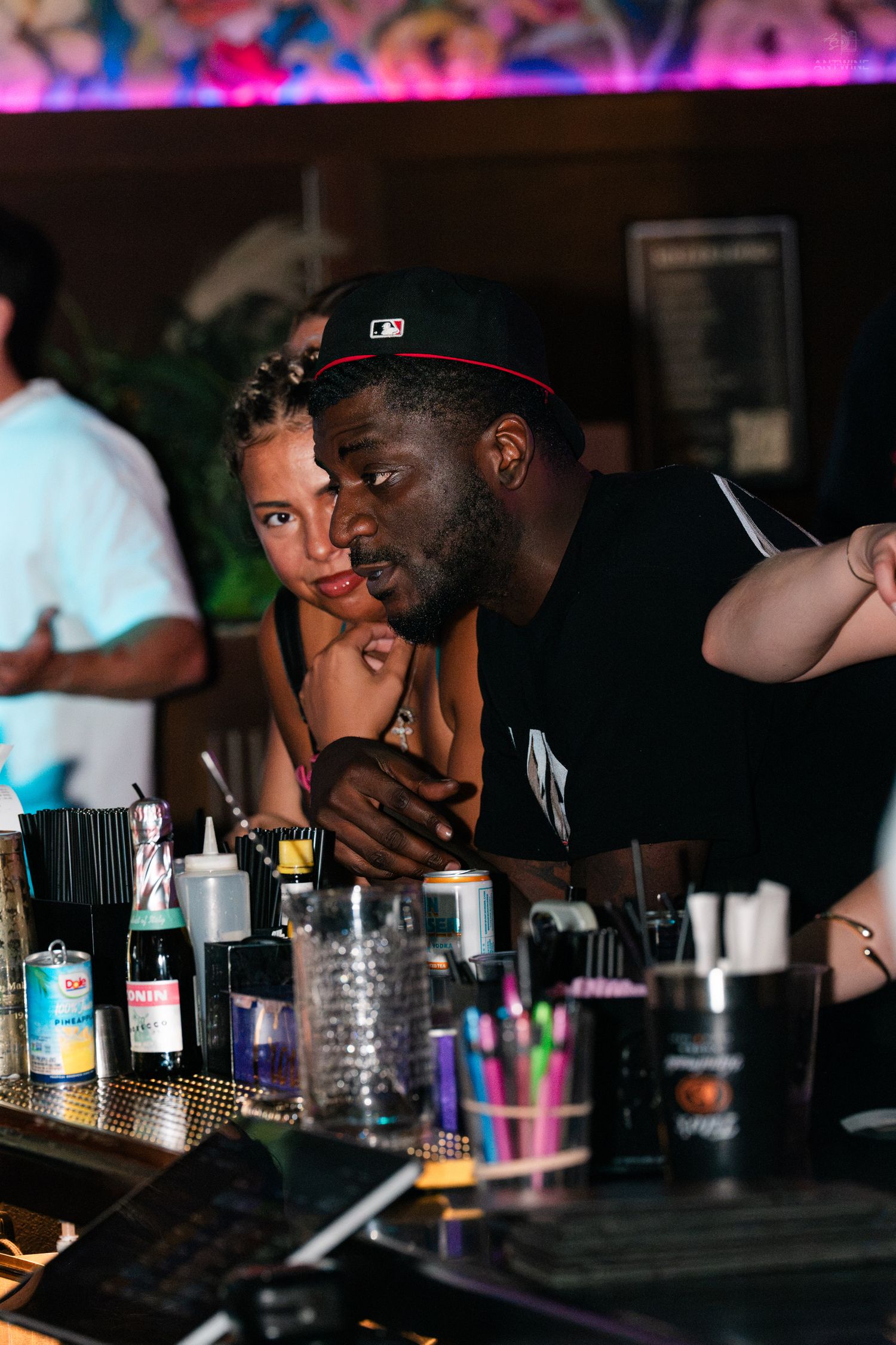 A man in a black MLB cap and woman lean over a bar counter filled with colorful supplies at a nightclub.