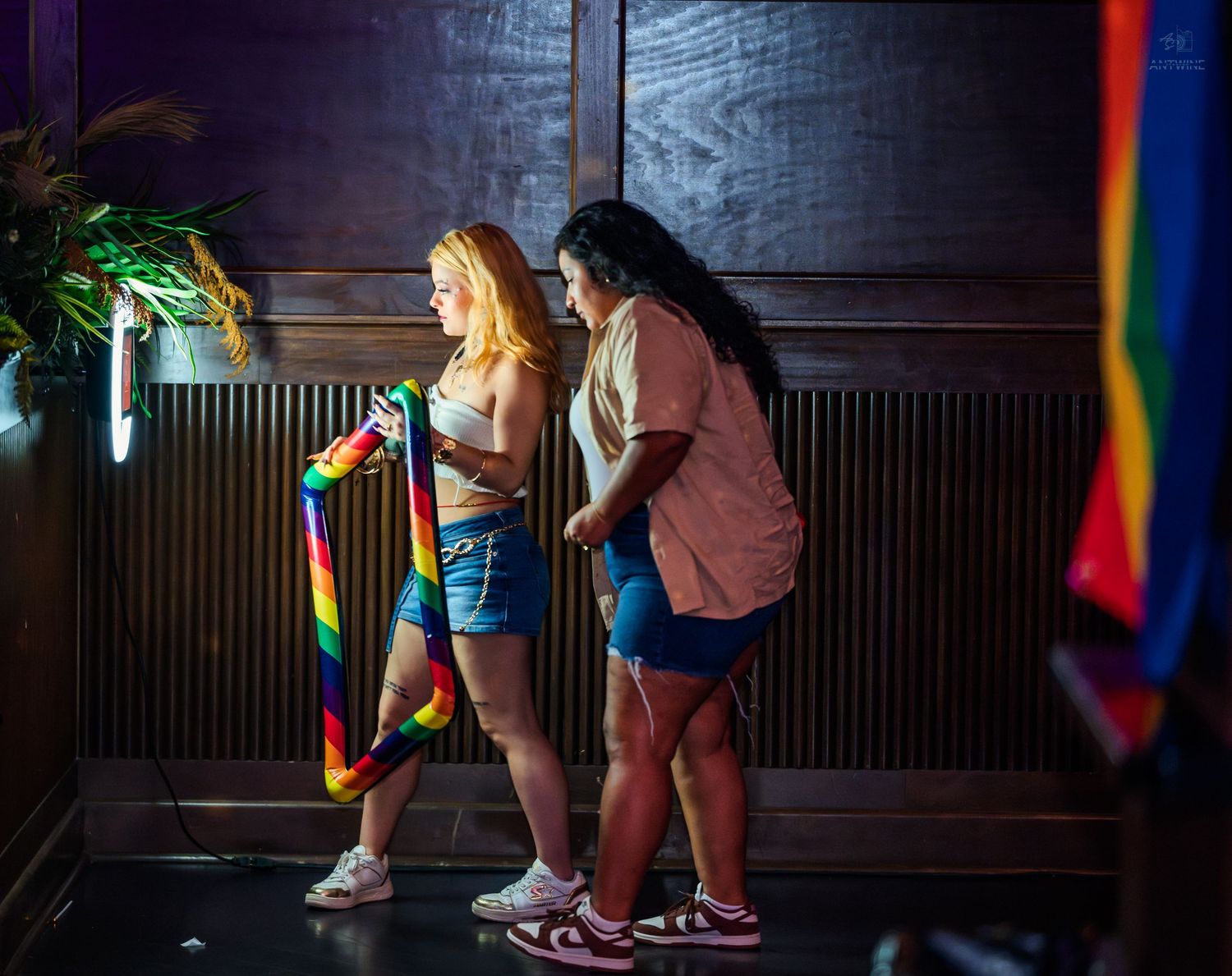 Two women holding a rainbow Pride flag in a dimly lit wooden interior venue.