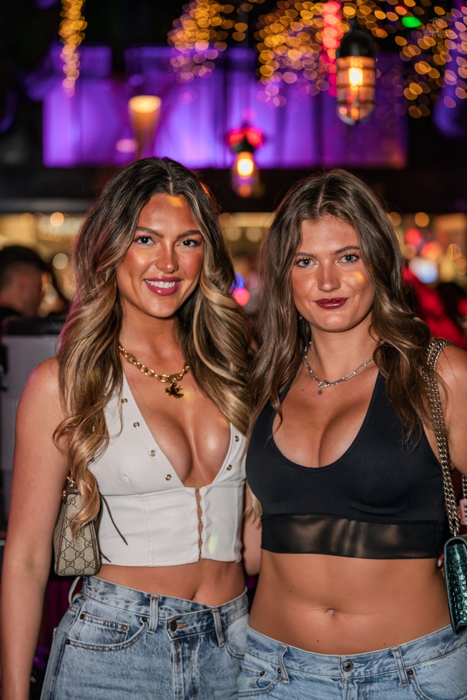 Two young women in trendy outfits smiling together at a nightclub with warm ambient lighting.