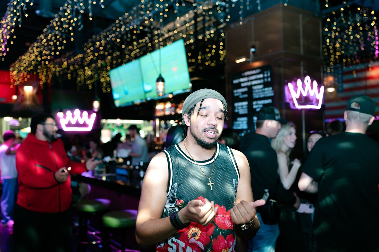 Man in mesh top holding flowers poses near a neon-lit bar with crown signs.