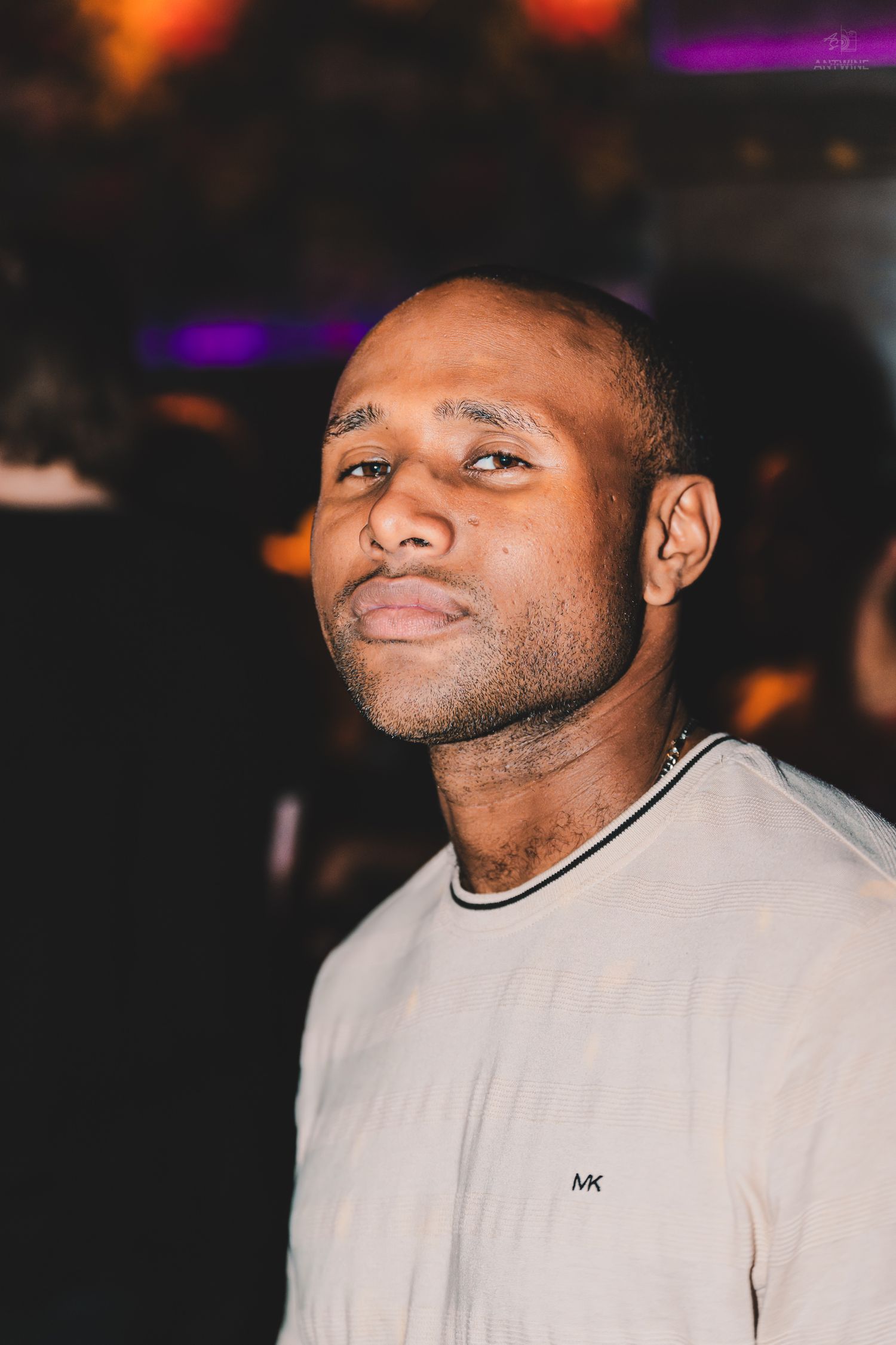 A bald Black man in a Michael Kors cream t-shirt gazes upward in a dimly lit nightclub.