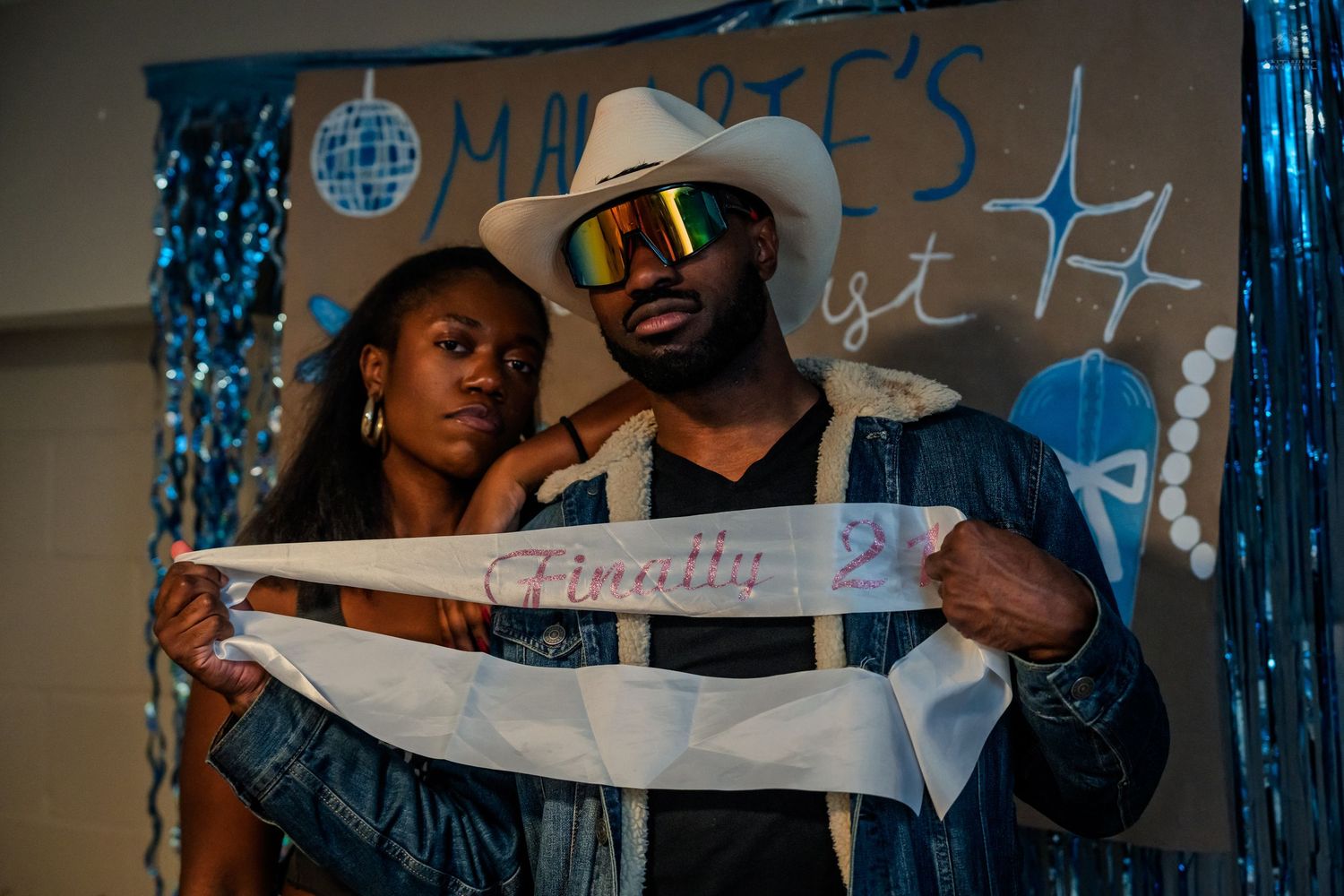 Couple posing together holding a 'Finally 21' sash at a blue-themed birthday party celebration.