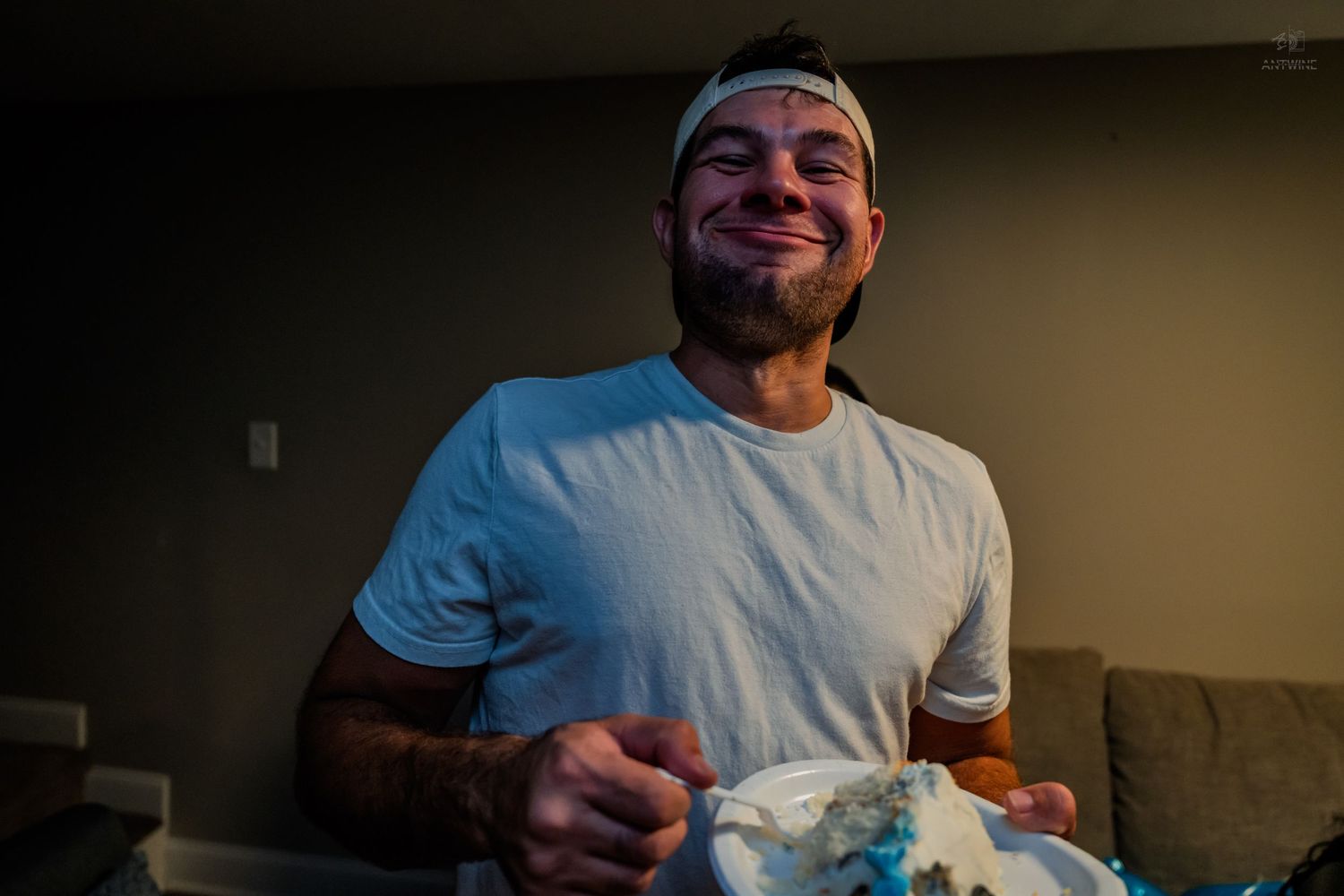 Man laughing with frosting on his face while holding a piece of birthday cake at a party.