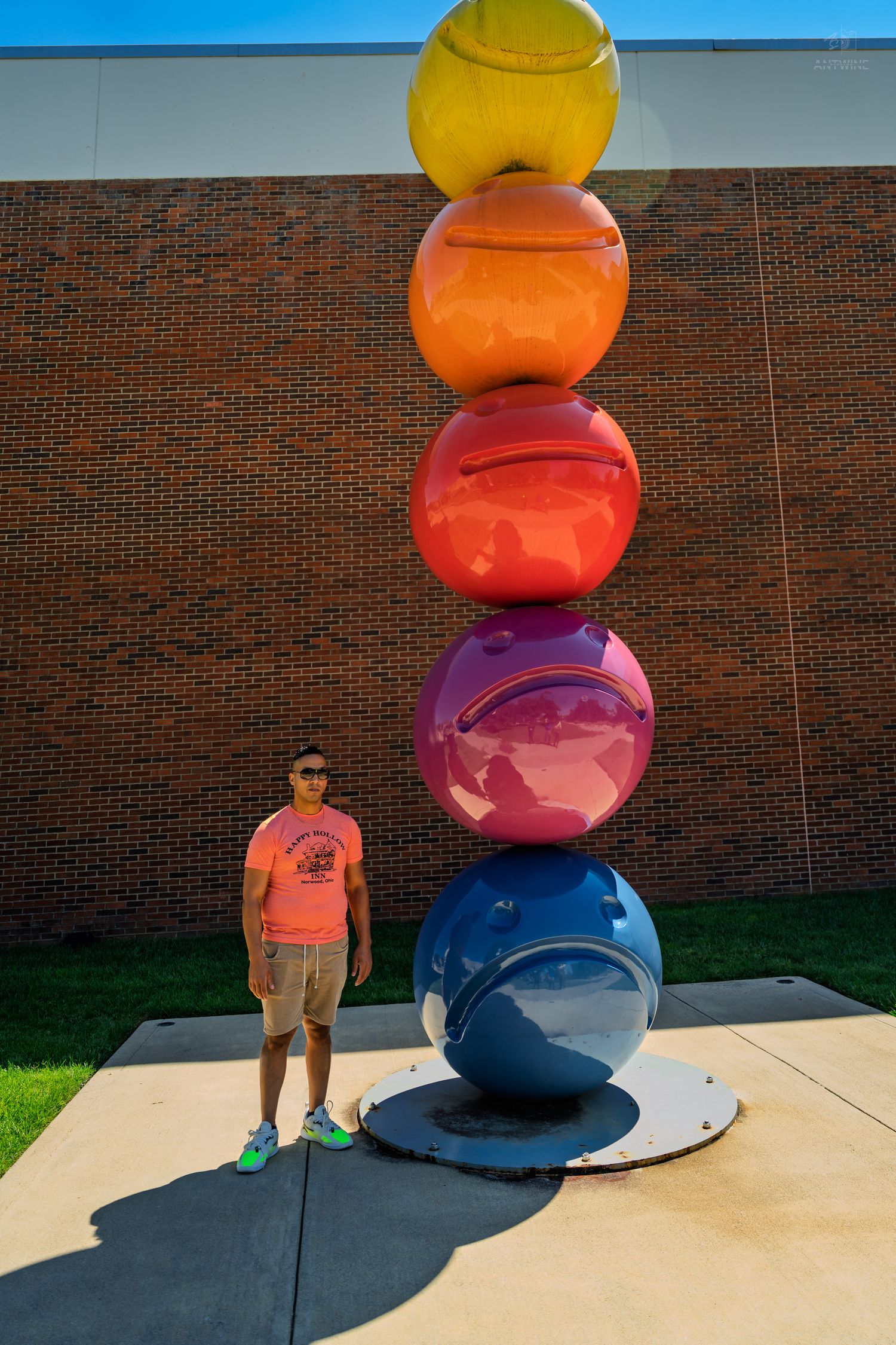 A man in an orange shirt poses next to a towering colorful stacked spheres outdoor sculpture.