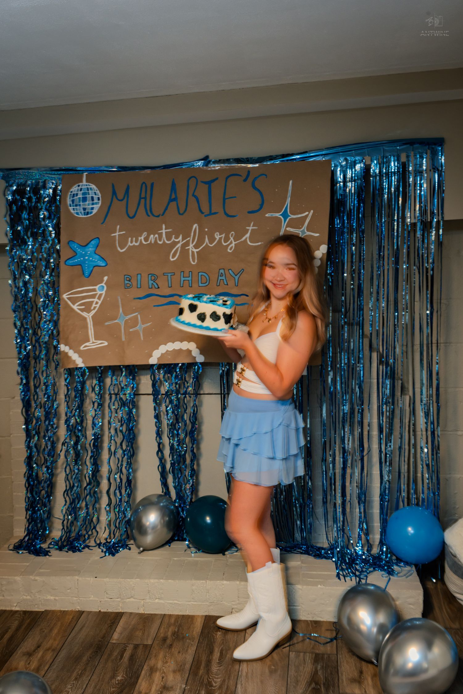 Woman in white crop top and blue ruffle skirt holds heart-shaped birthday cake at Malarie's 21st birthday celebration.