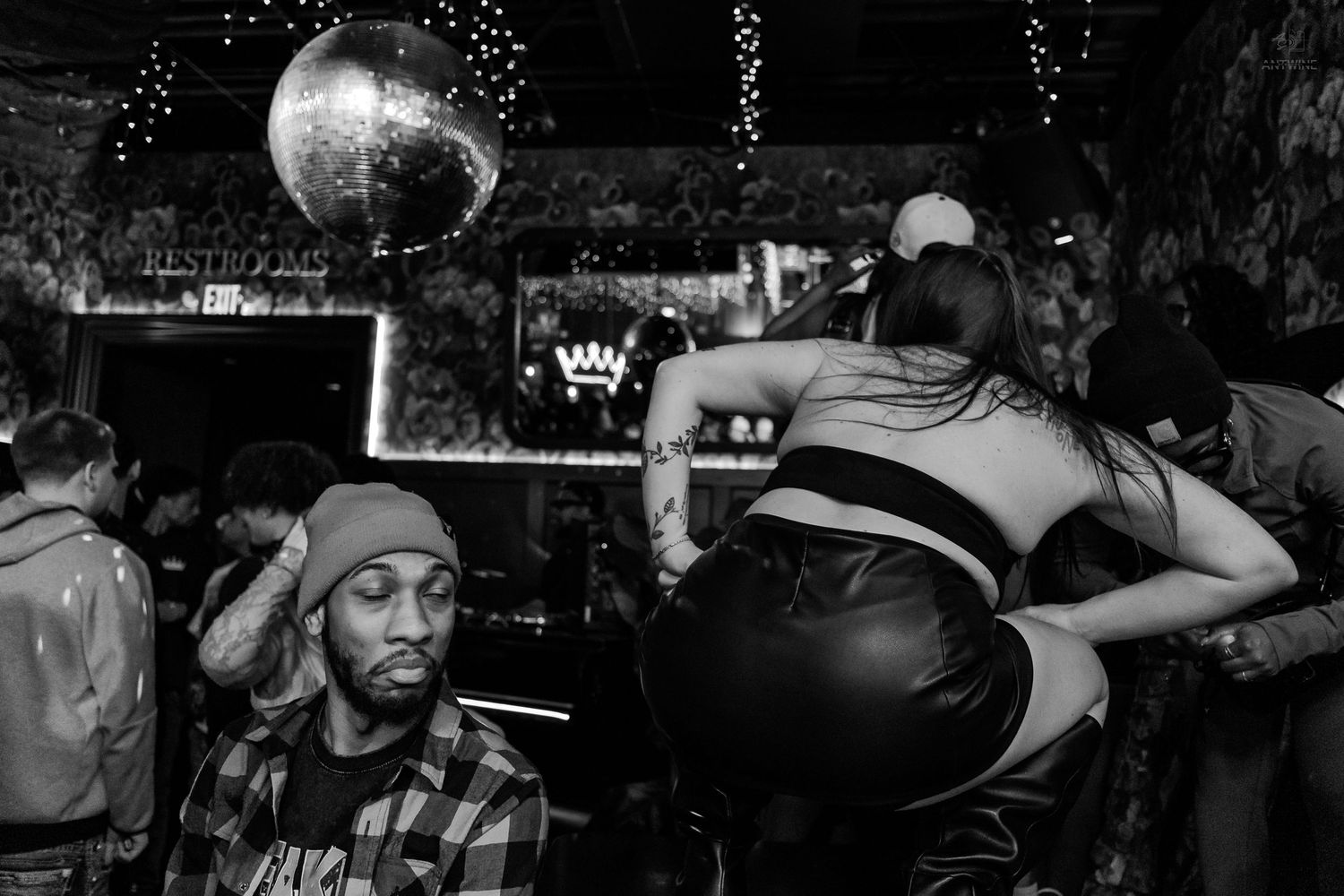 Man in plaid shirt sits near disco ball as people dance at nightclub event.