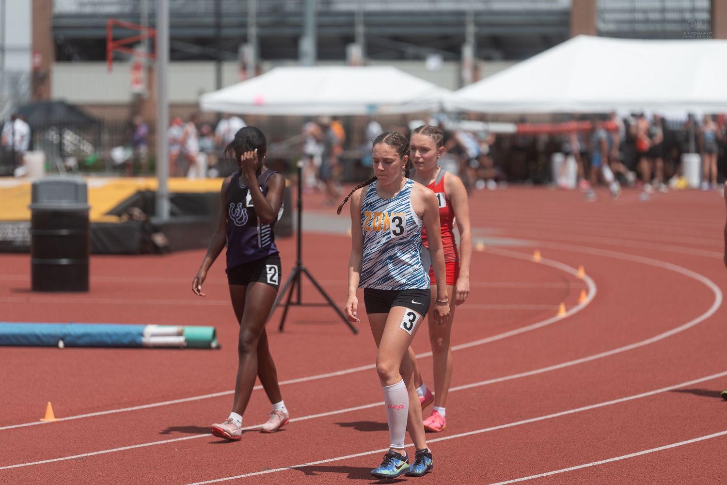 Three female track runners in competition uniforms walking on red track after finishing a race outdoors.