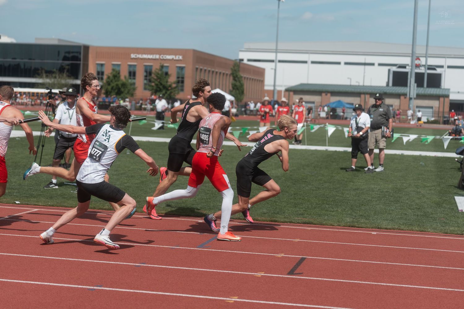 Young male athletes sprint on a red track during a high school relay race competition on a sunny day.