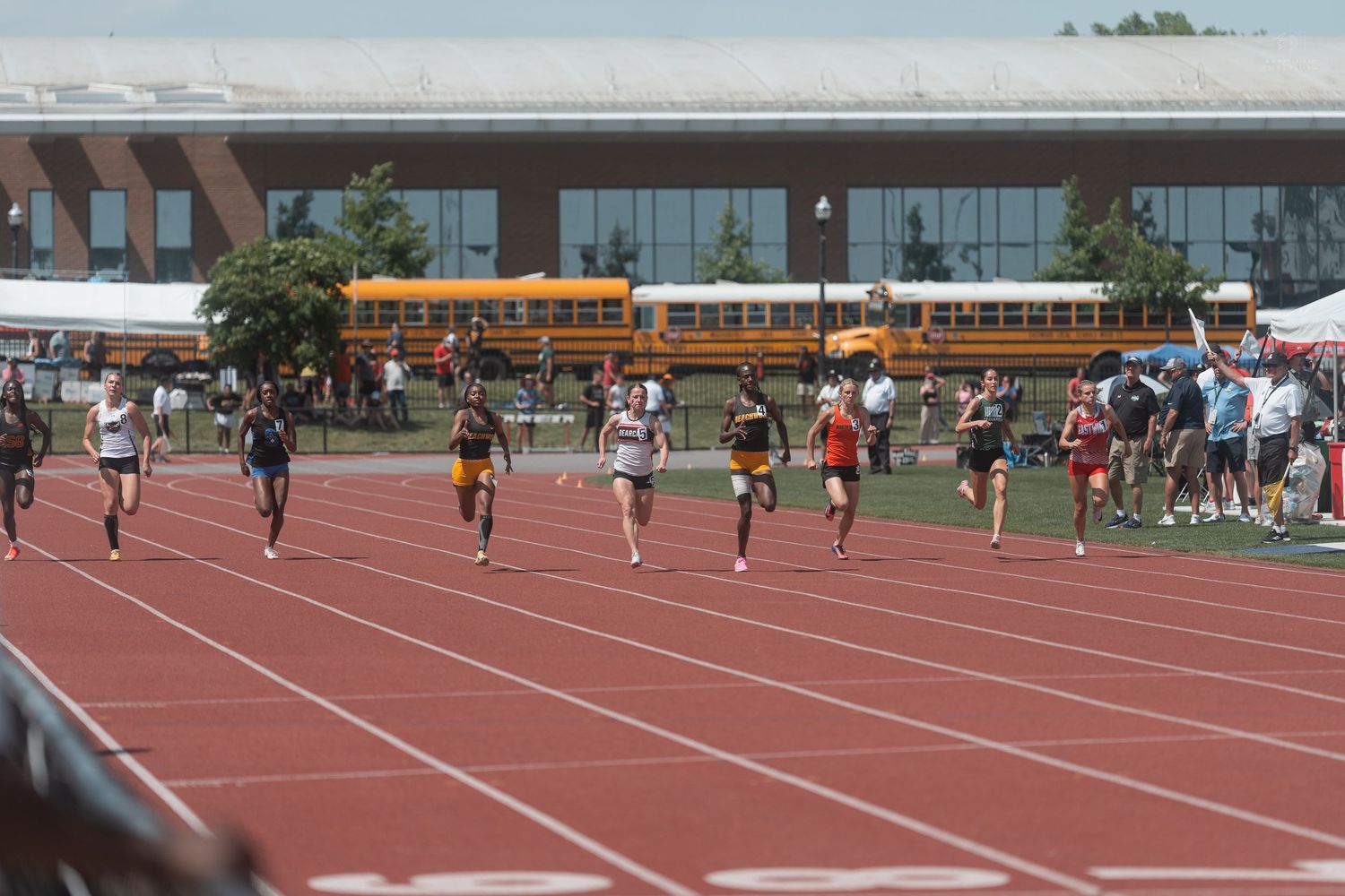 High school track athletes compete in a sprint race on a red track with yellow school buses in the background.