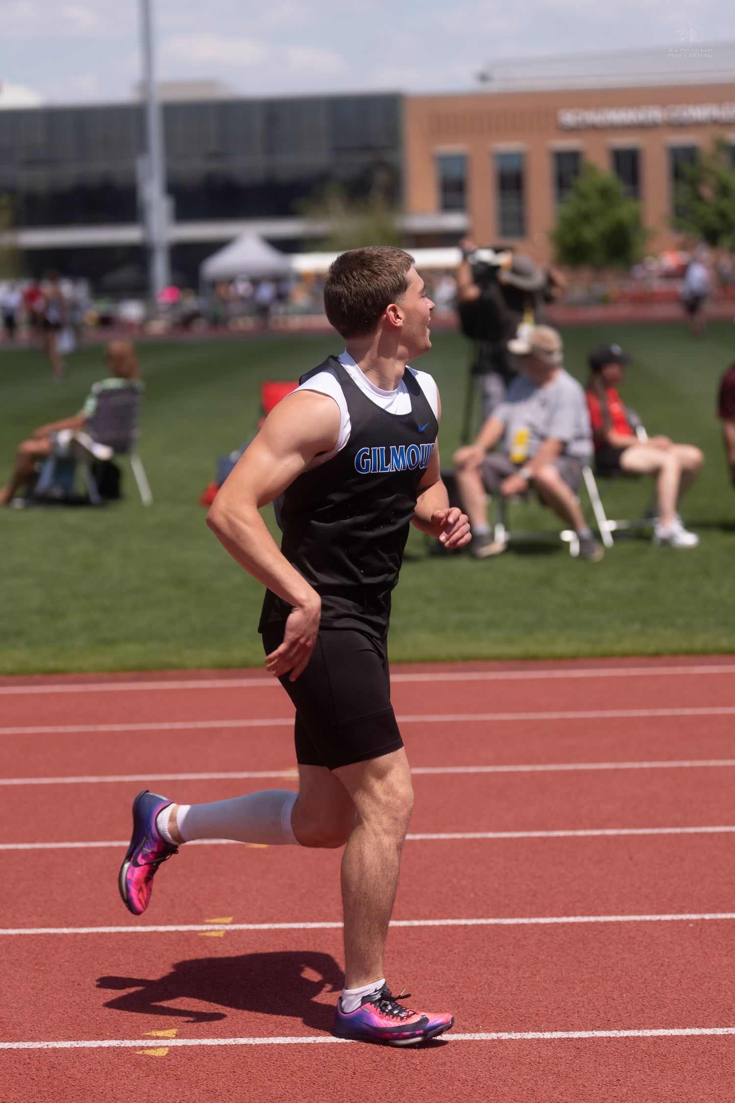 Male athlete wearing a Gilmore jersey runs on a red track during an outdoor track and field competition on a sunny day.
