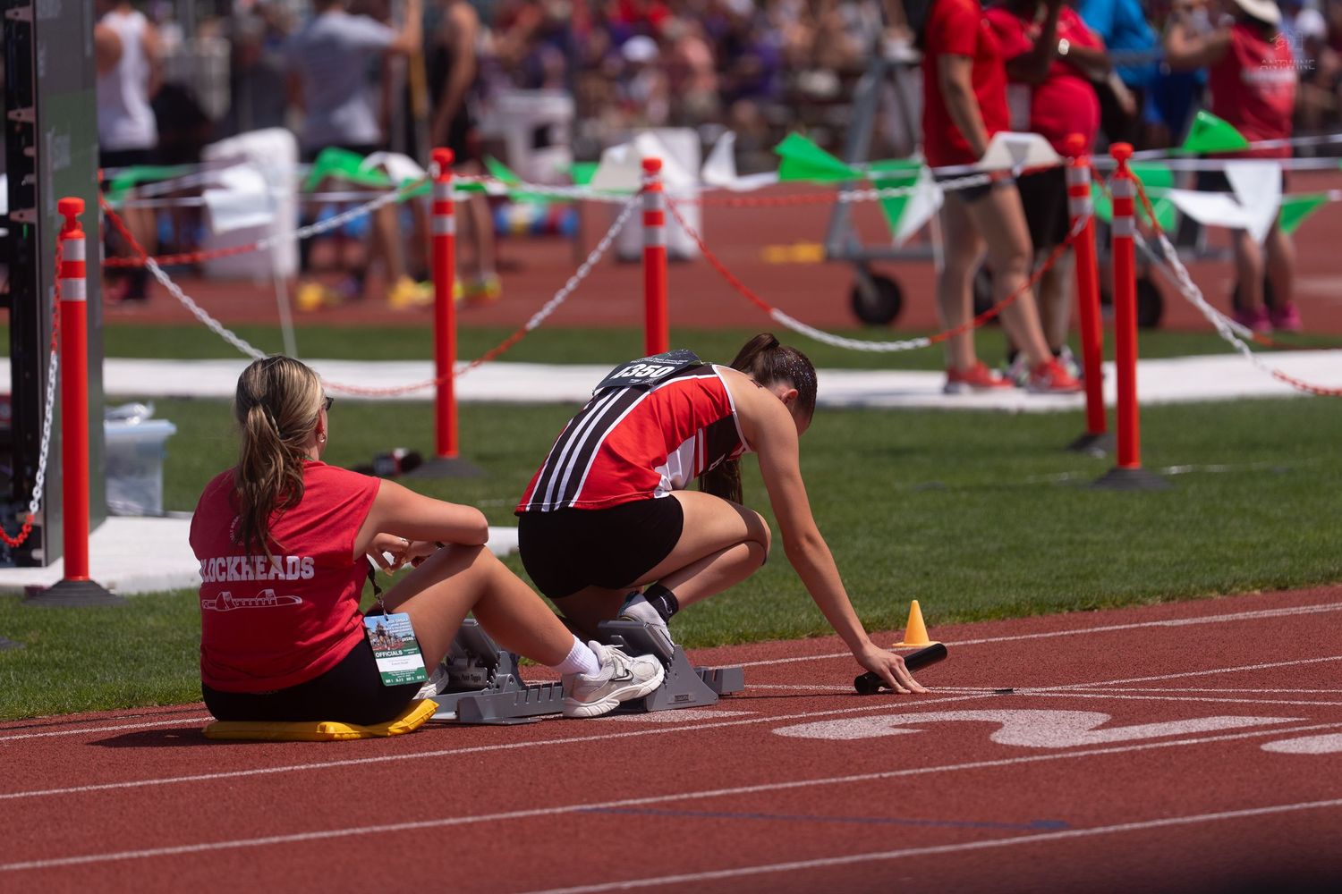 Two female track athletes prepare in starting blocks on a red track at an outdoor athletic competition.