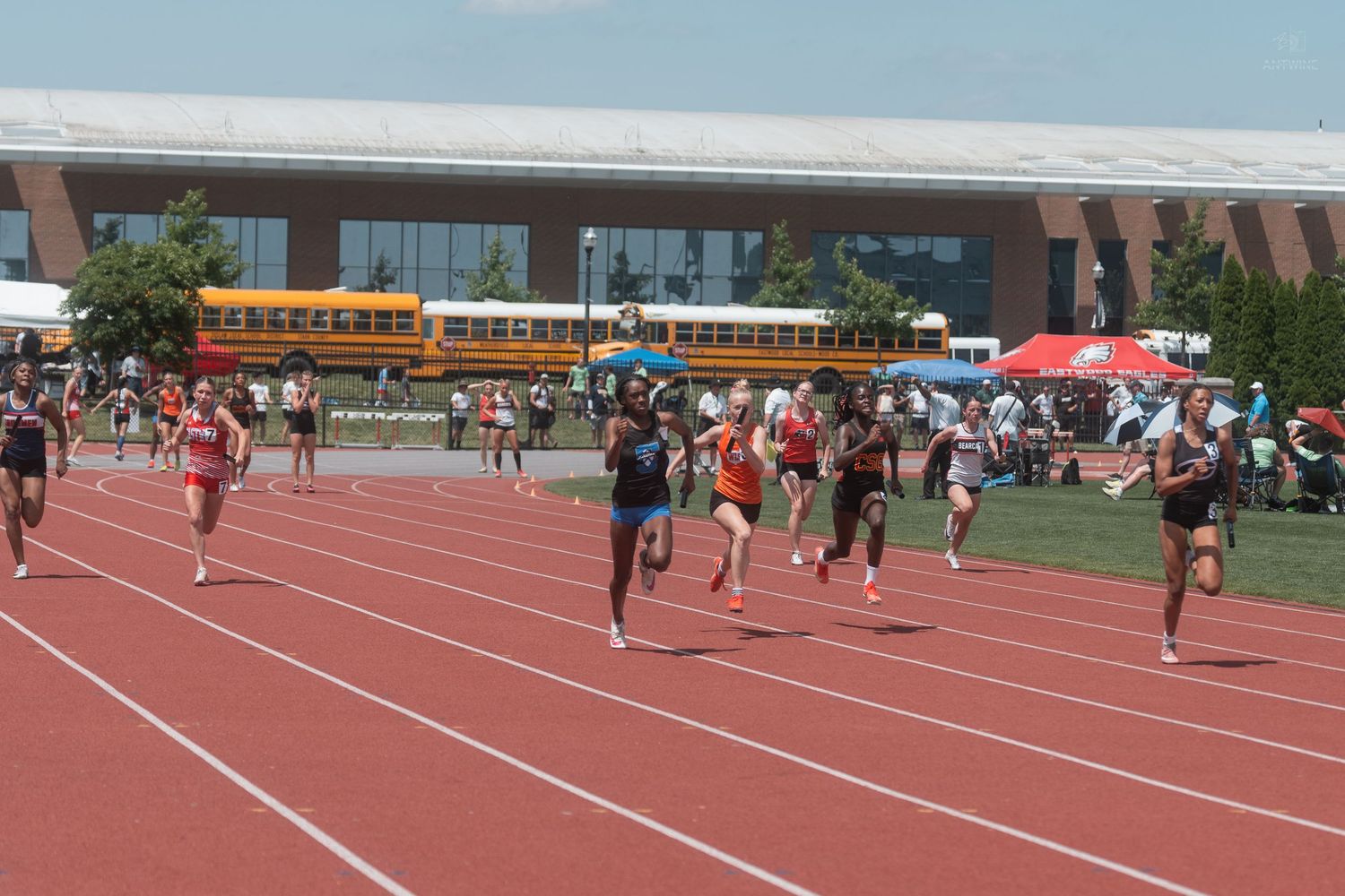Runners competing in sprint race on red track at outdoor collegiate track and field meet on sunny day.