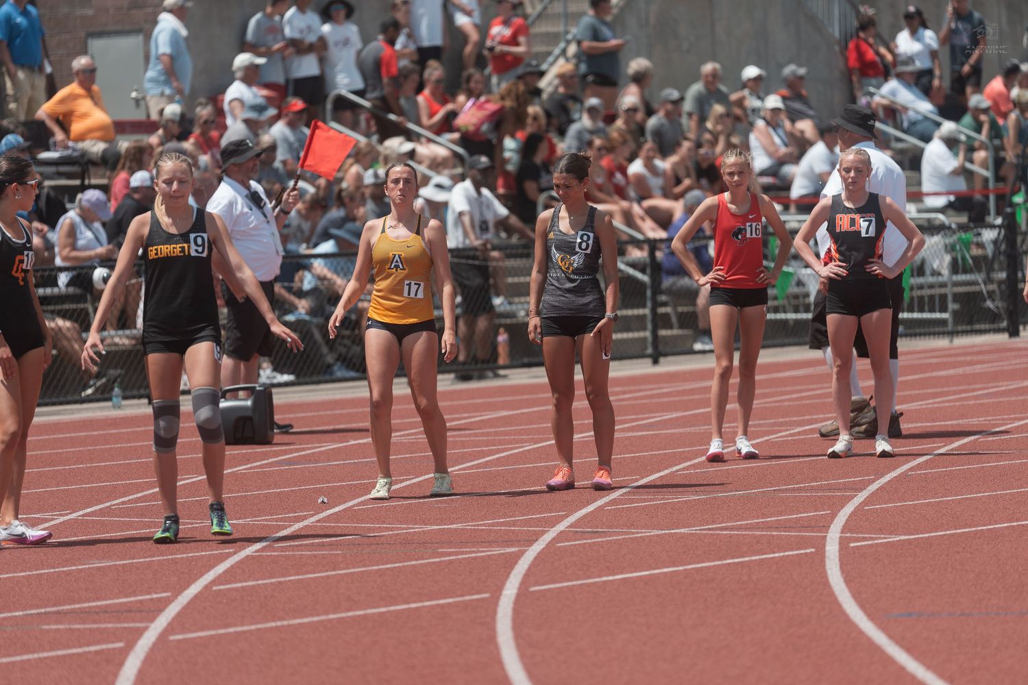 Female track athletes prepare to start a relay race on a red track at a crowded high school track and field meet.