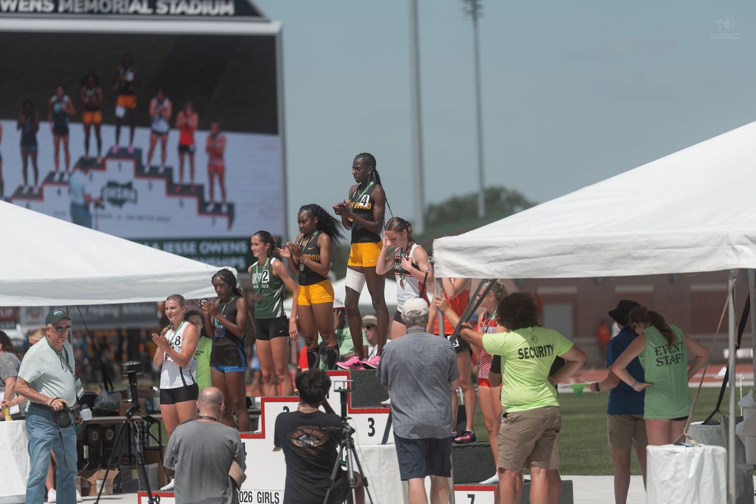 Young female athletes receive medals on the awards podium at an outdoor track and field championship event.