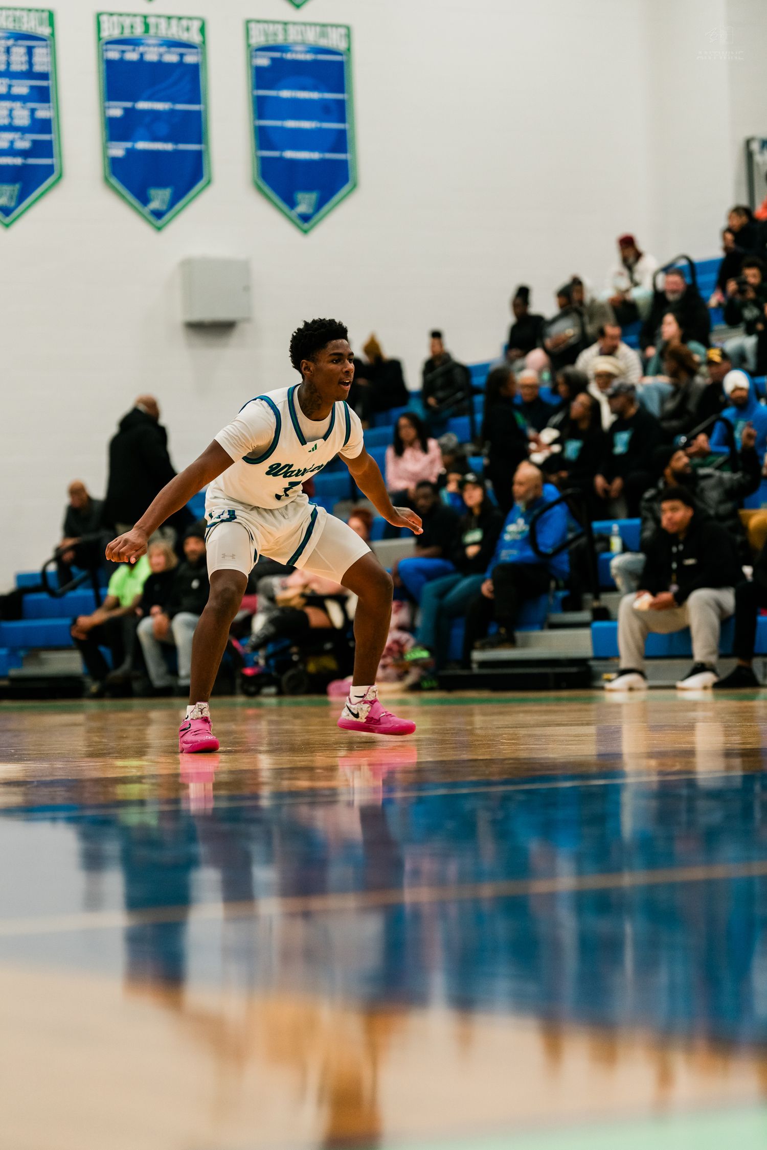 Warriors basketball player in white uniform takes defensive stance on shiny court before packed gymnasium crowd.