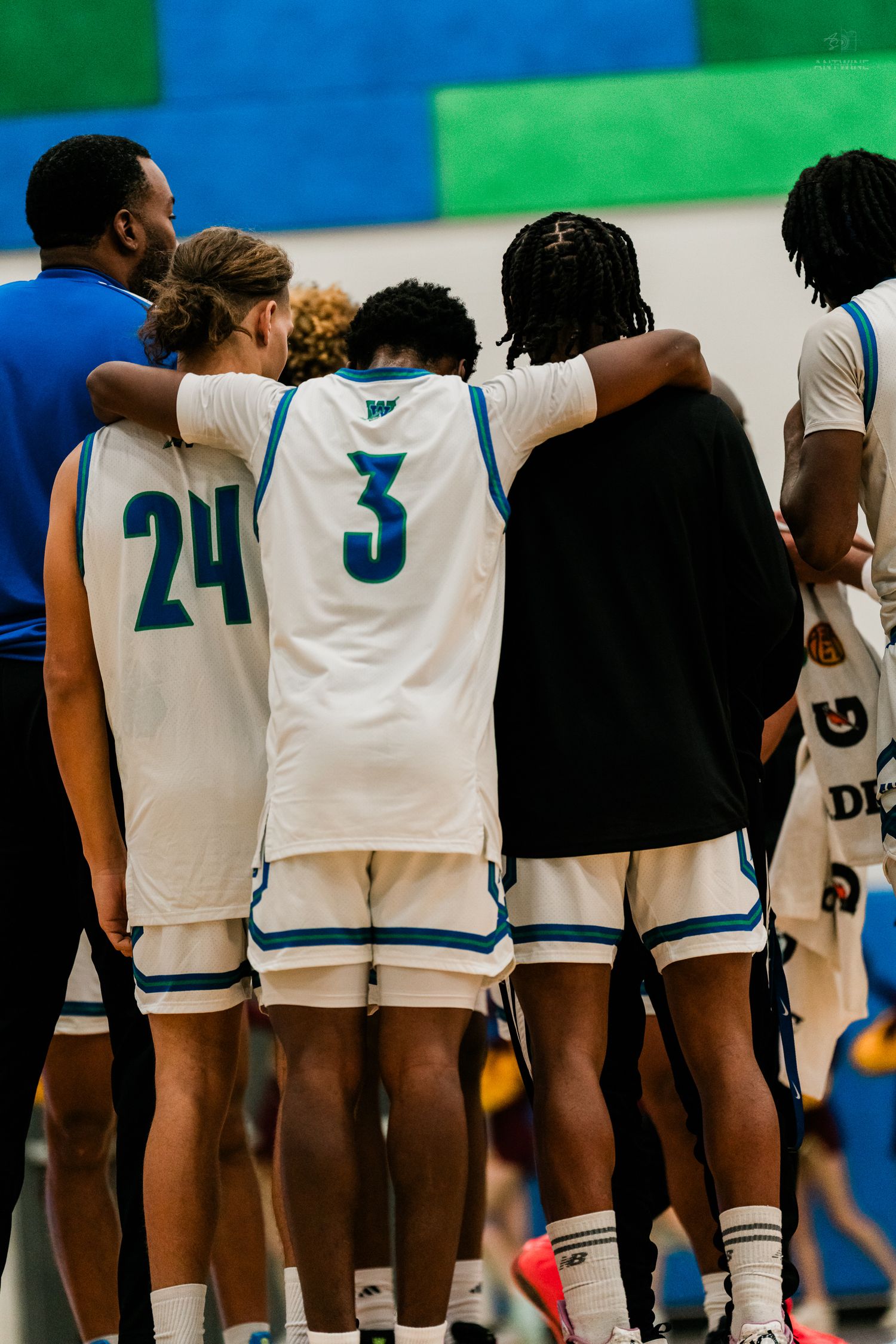 High school basketball players #24 and #3 in white Warriors uniforms huddle with teammates during a timeout.