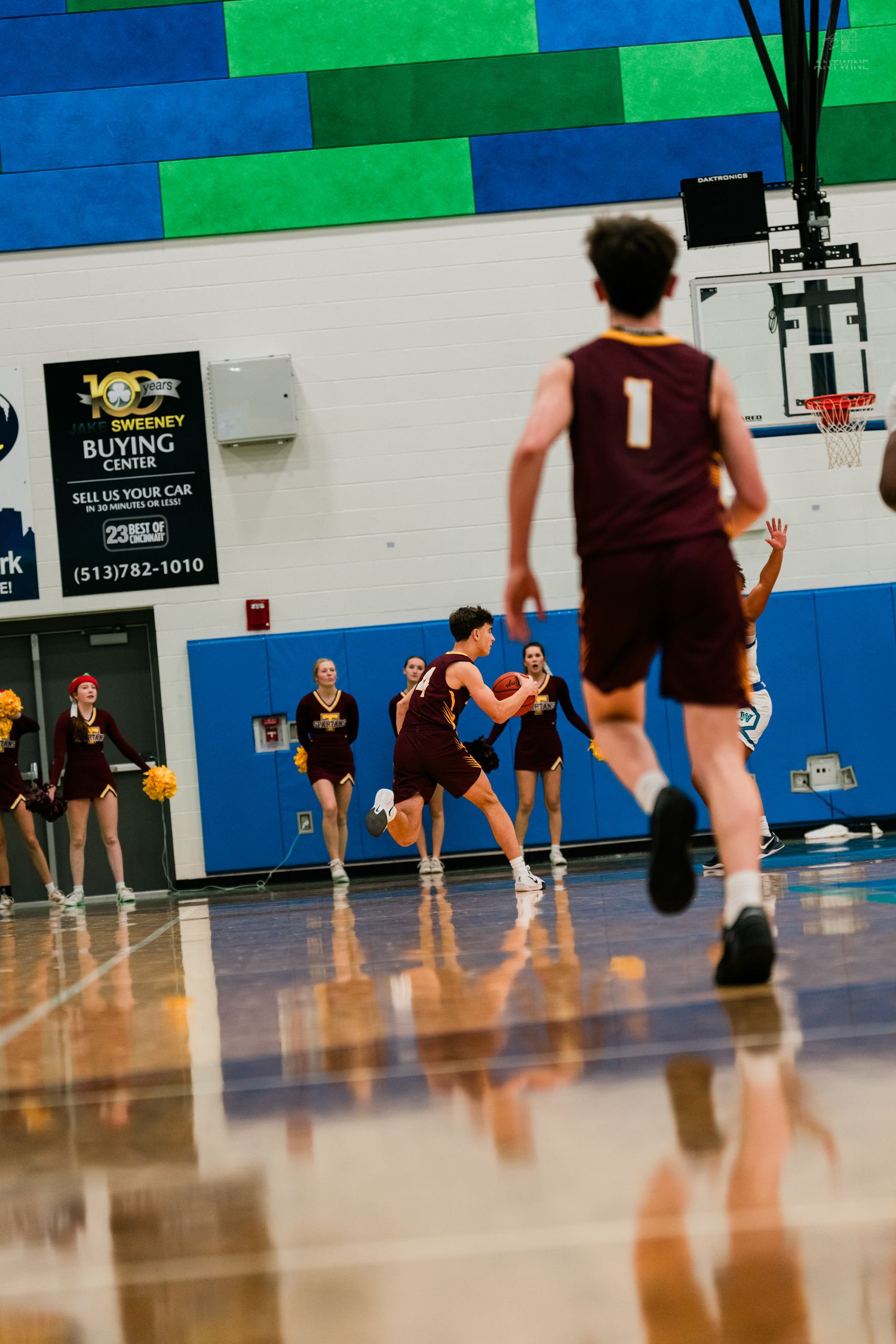 Turpin High School basketball players in maroon uniforms dribble during a varsity game in a blue-walled gymnasium.