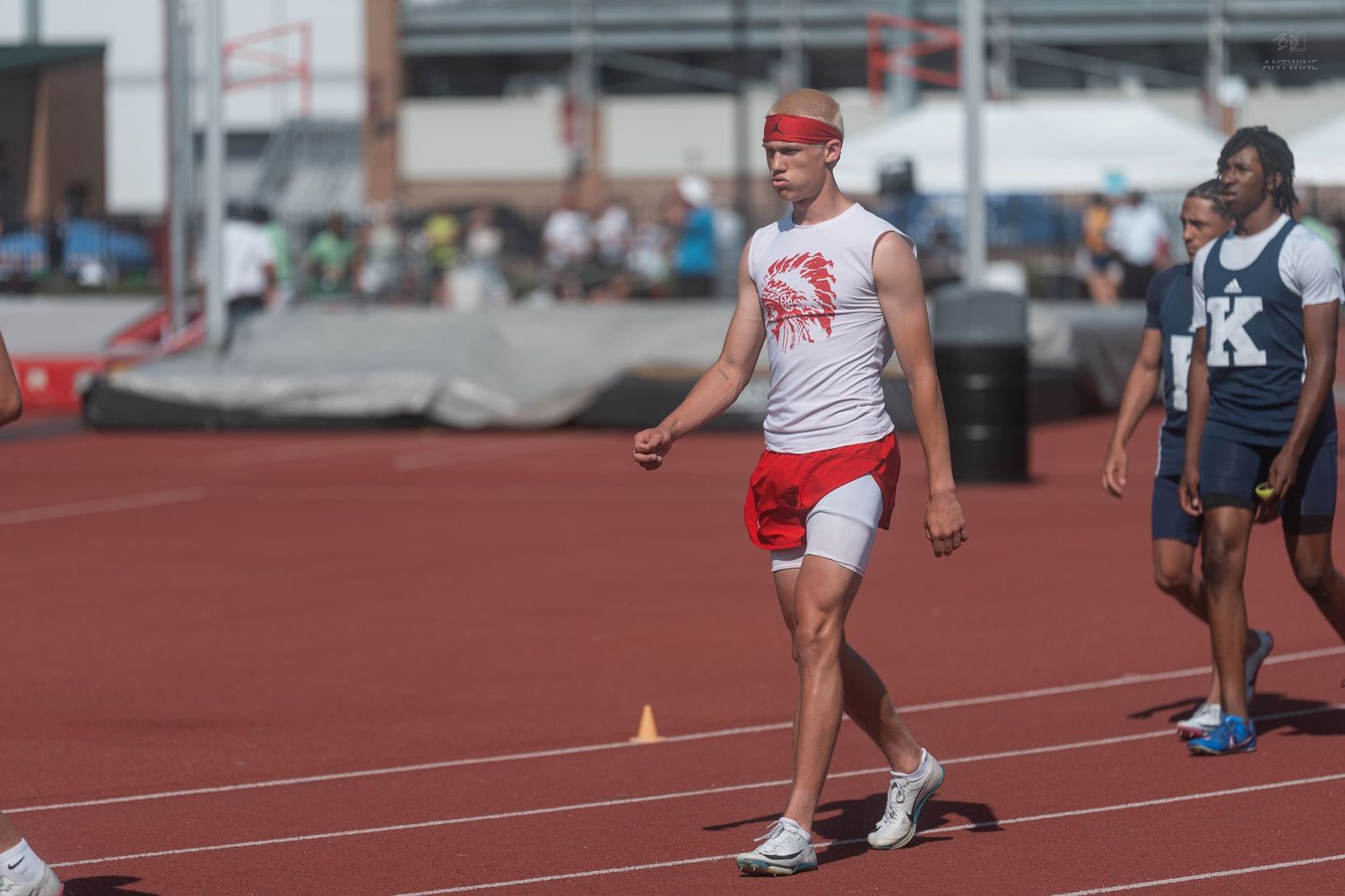 Athlete in red and white uniform with headband walks on track during outdoor track and field competition.