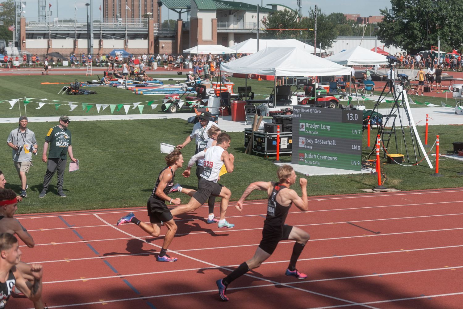 High school sprinters race down a red track at an outdoor track and field meet on a sunny day.