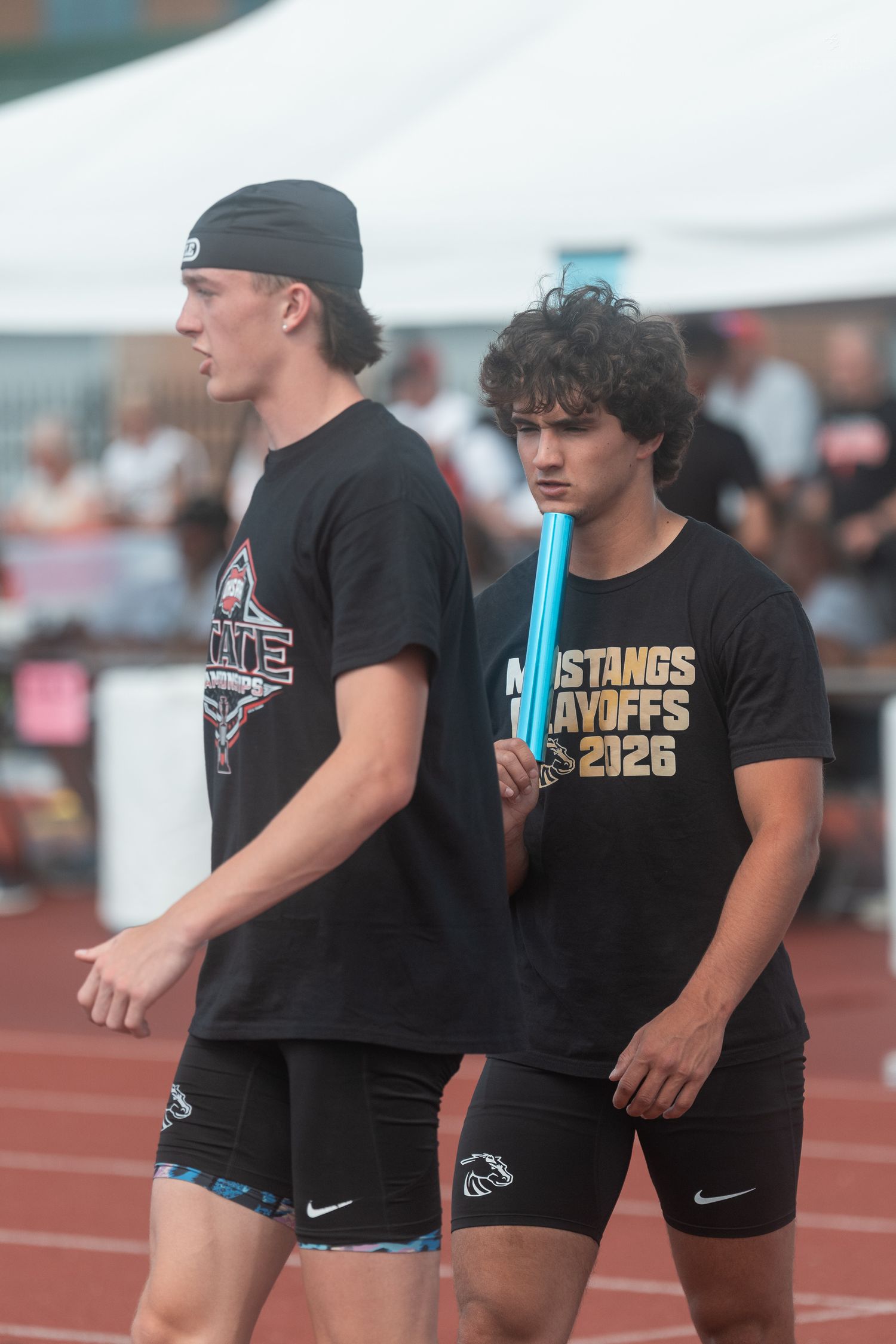 Two male track athletes in black Nike uniforms stand on a red track, one holding a blue relay baton at a playoff meet.