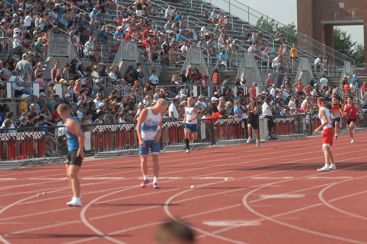 Track and field athletes prepare on a red running track with a large crowd filling the stadium bleachers behind them.