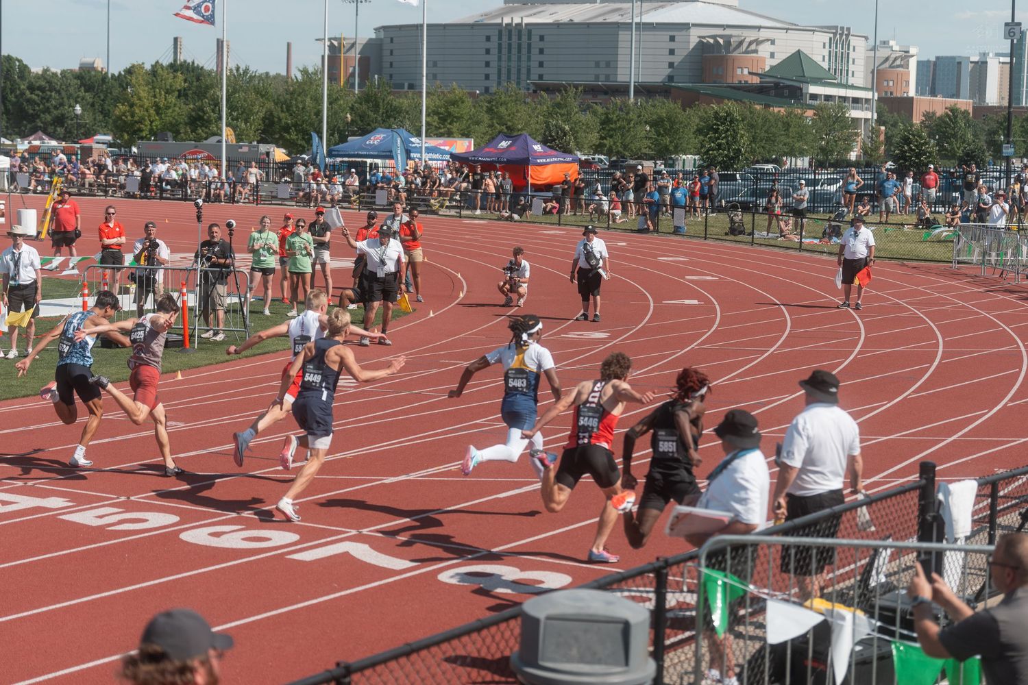 Athletes sprint across a red track during a competitive outdoor track meet with crowds of spectators watching.