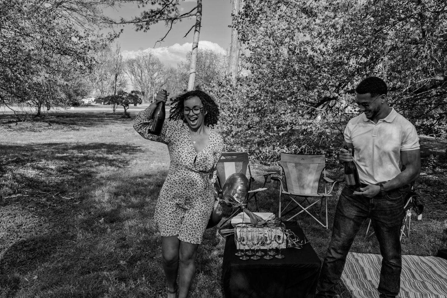 Black and white photo of couple laughing together at decorated outdoor picnic table under trees.
