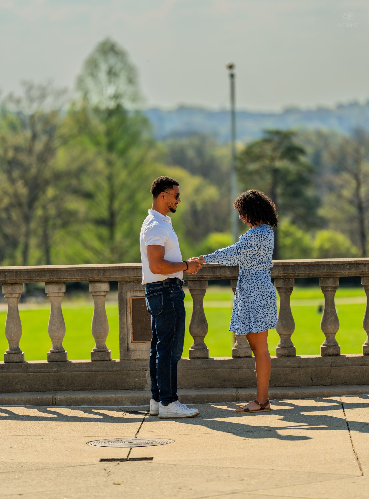 Couple holding hands facing each other on a sunlit stone terrace with green park views.
