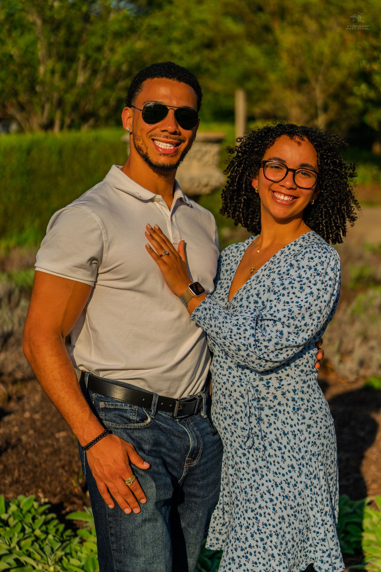 Smiling couple poses outdoors in golden hour light, woman in floral dress shows engagement ring.