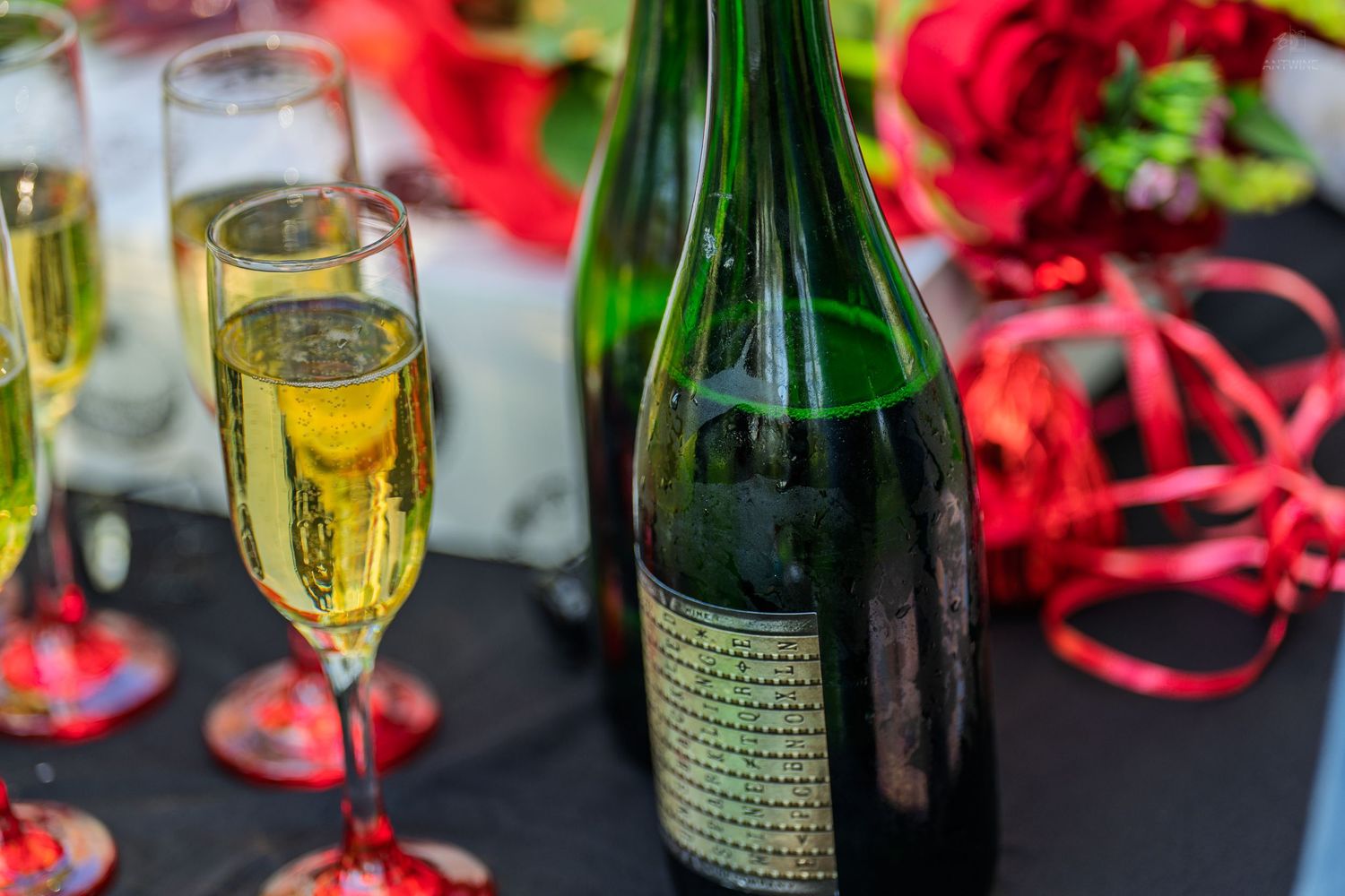 Champagne bottle and filled flutes on a black table with red roses and ribbons.